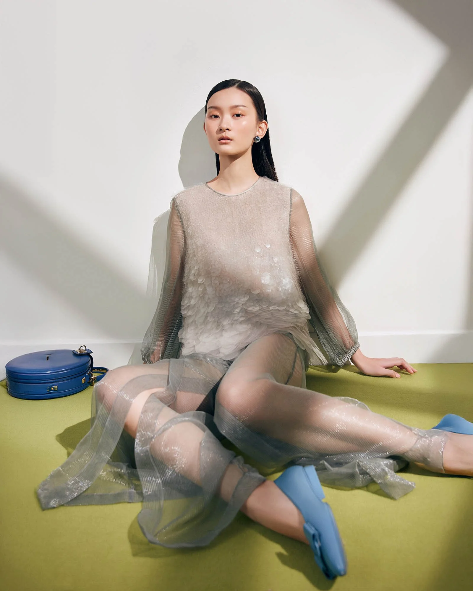 A woman sitting on a green floor against a white wall, wearing a sheer, textured dress with a blue bag nearby.
