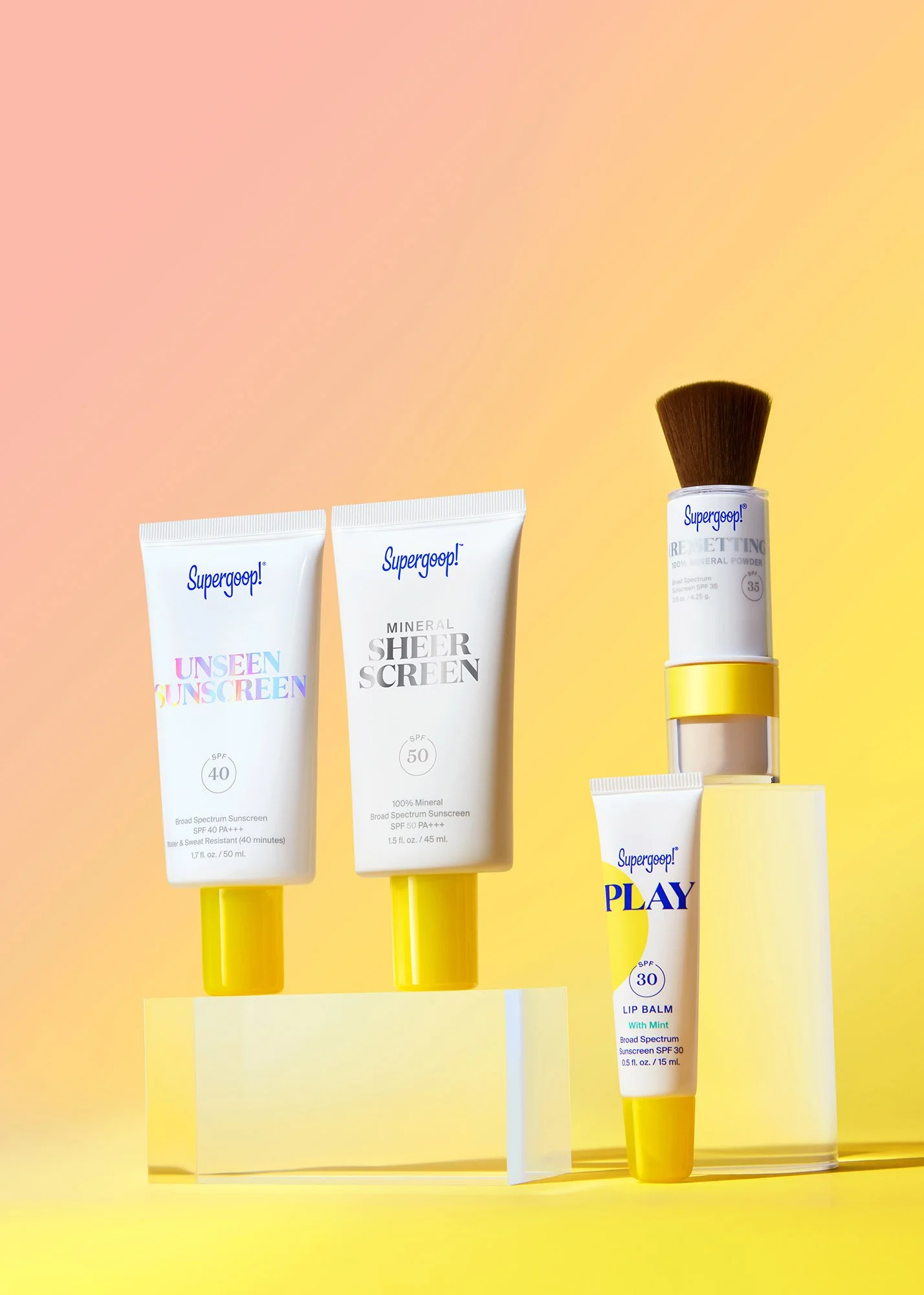 Display of five Supergoop! sunscreen and skincare products with yellow and pink gradient background. There are two tubes of sunscreen labeled "Unseen Sunscreen" SPF 40 and "Mineral Sheer Screen" SPF 50, a liquid foundation or concealer with a brush a
