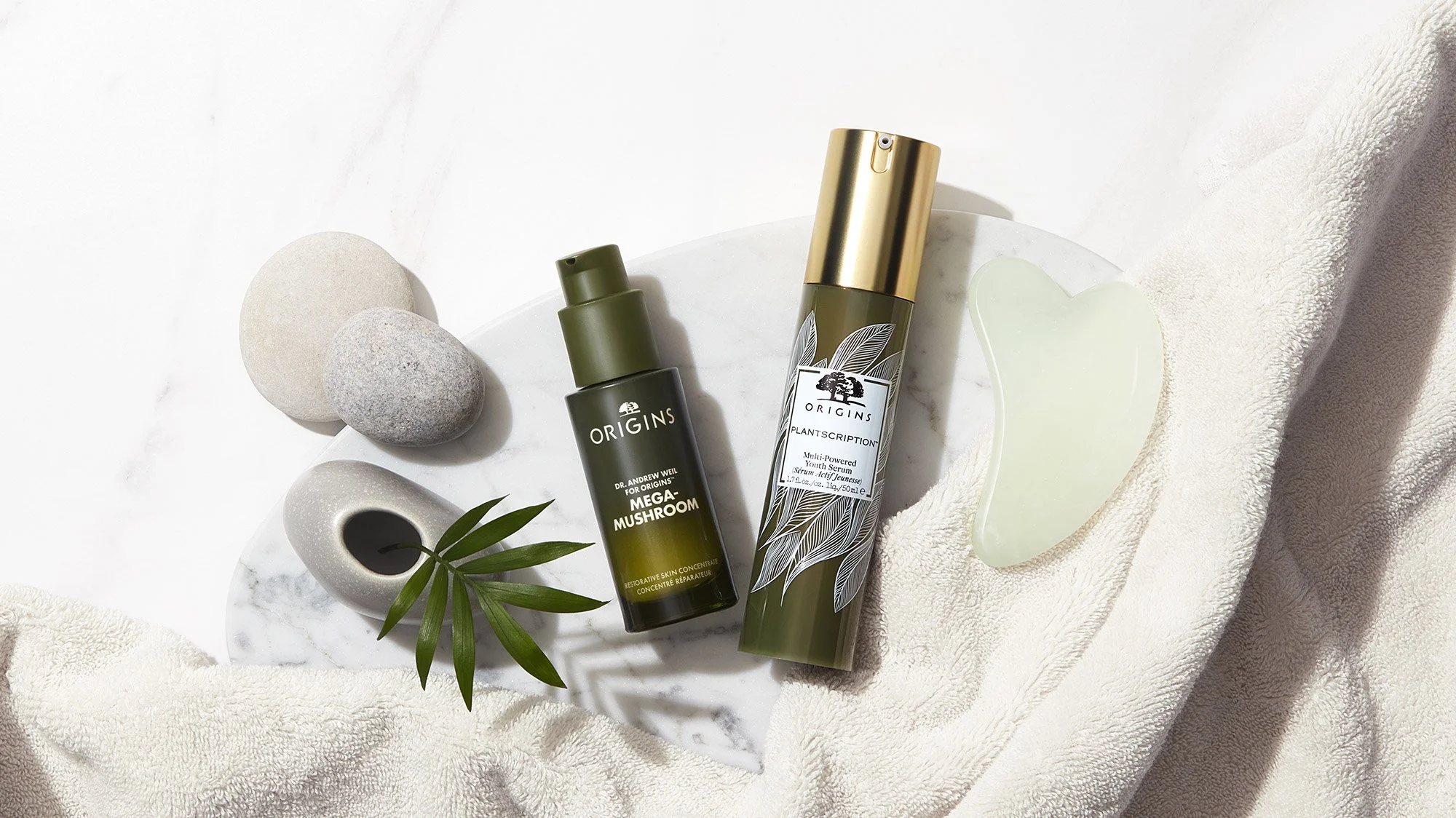 Skincare products on a white marble tray, including a green bottle of Origins Mega-Mushroom serum, a taller bottle of Origins Plantscription youth serum, surrounded by smooth stones, a small white vase with green leaves, a jade roller, and a beige to