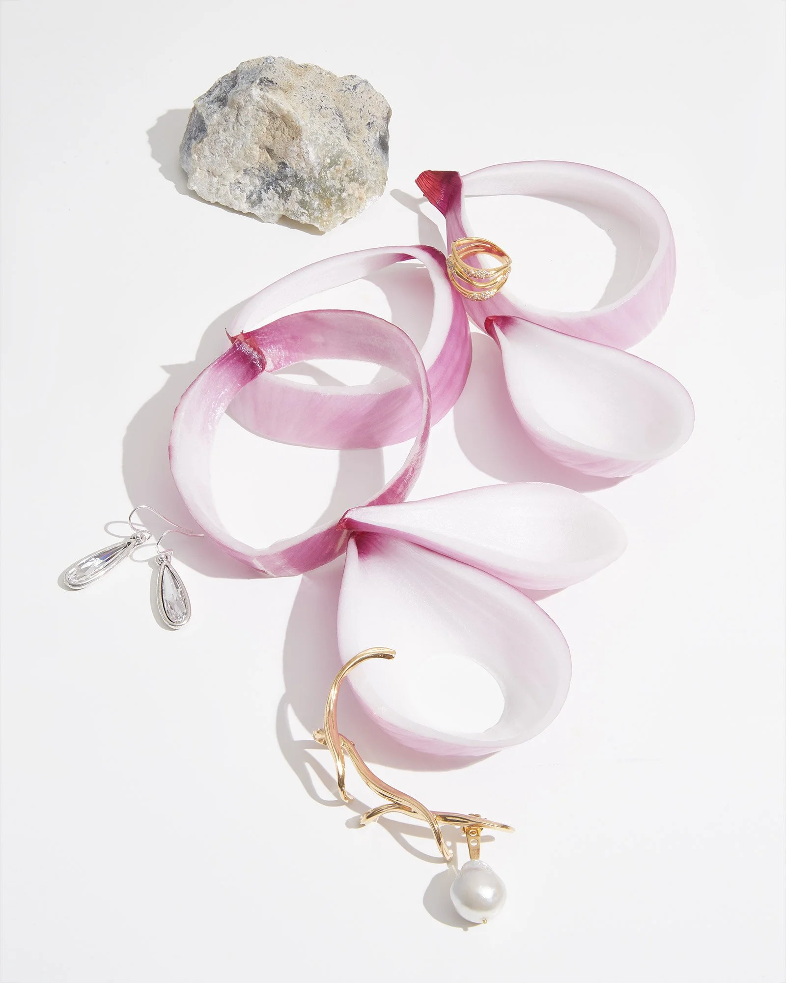 Pink and white flower petals, a gray rock, and gold, silver, and pearl jewelry arranged on a white surface.