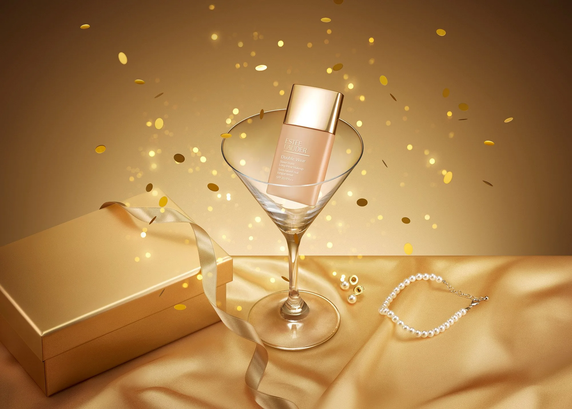 A gold-colored gift box with a gold ribbon, a pearl necklace, and a pearl bracelet are on a silky gold fabric. A large martini glass contains a bottle of Estée Lauder Double Wear makeup product, with gold and silver confetti falling around.