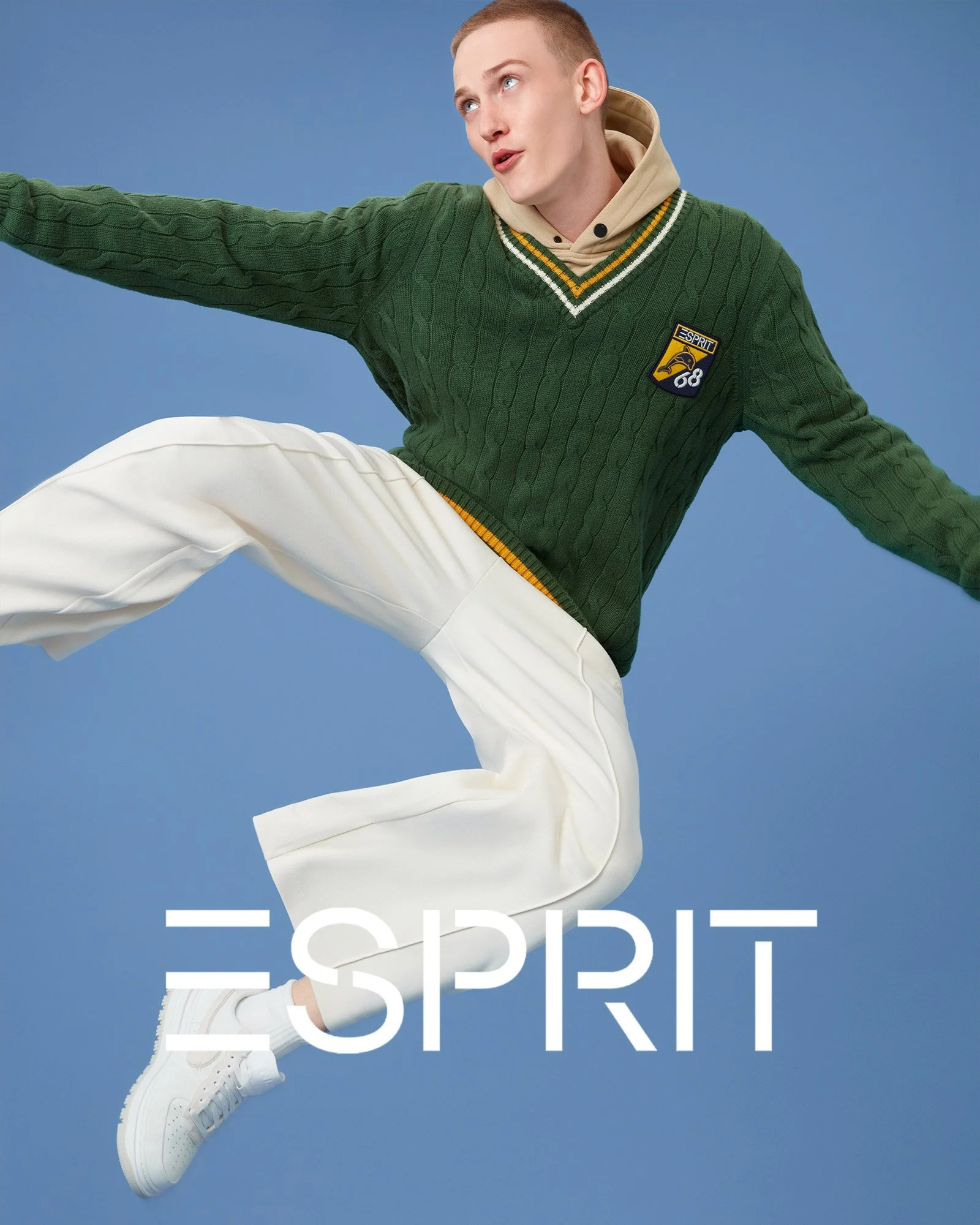 A young man in a green cable knit sweater with a 'Esprit' patch, beige hoodie, white pants, and white sneakers, jumping against a blue background with the 'Esprit' logo at the bottom.
