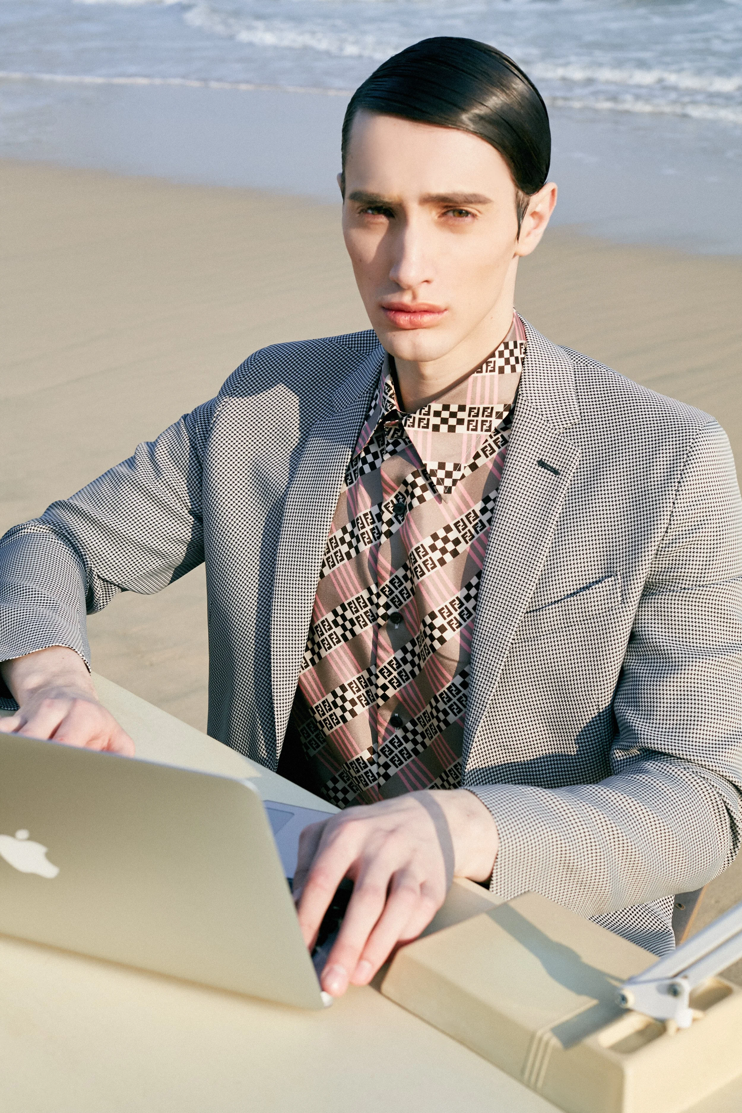 A young man with slicked-back black hair working on a laptop at the beach, dressed in a patterned blazer and shirt.