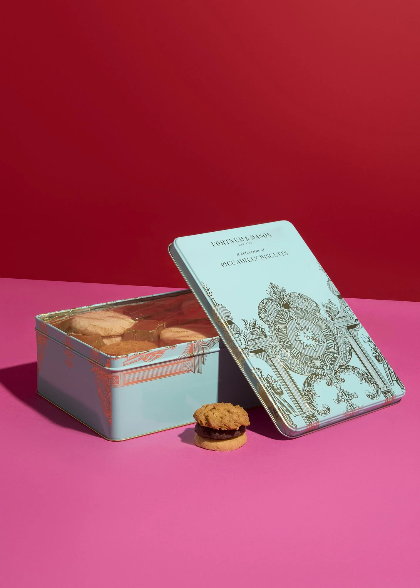 A blue decorative tin box with a selection of Piccadilly biscuits inside, with one biscuit and a cookie in front, on a pink surface against a red background.
