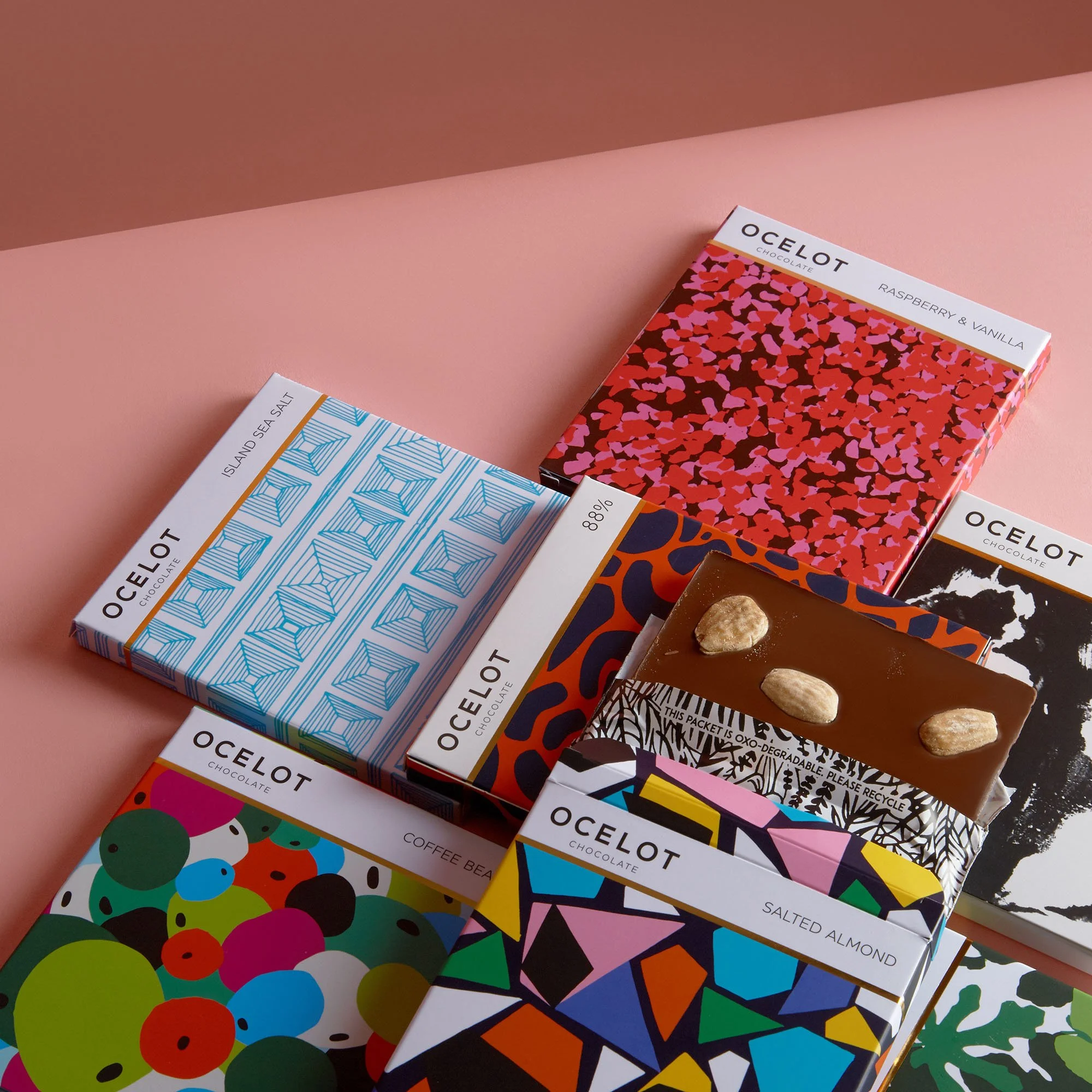 Several colorful chocolate bars with unique patterned packaging, some with almonds on top, placed on a pink surface.