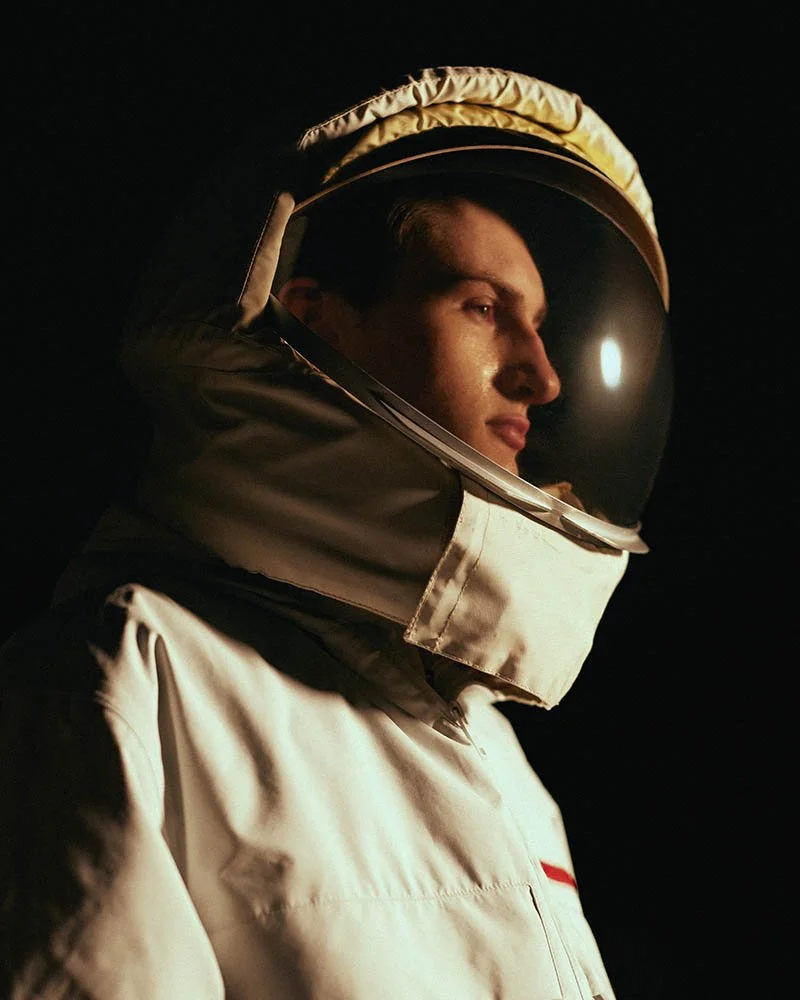 Person wearing a white space suit and helmet, looking to the right against a dark background.