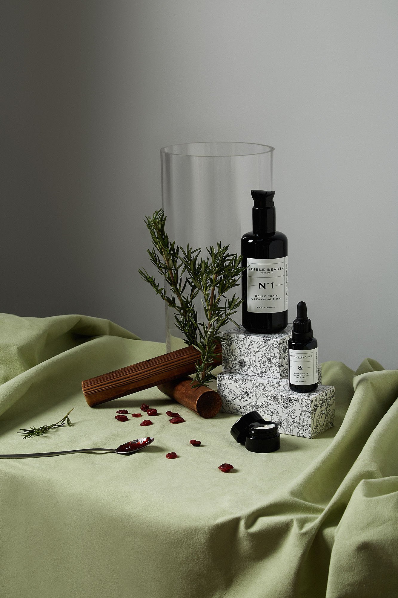 Skincare products on a table with green fabric, herbal sprig, wood sticks, and spilled red berries, set against a gray background.