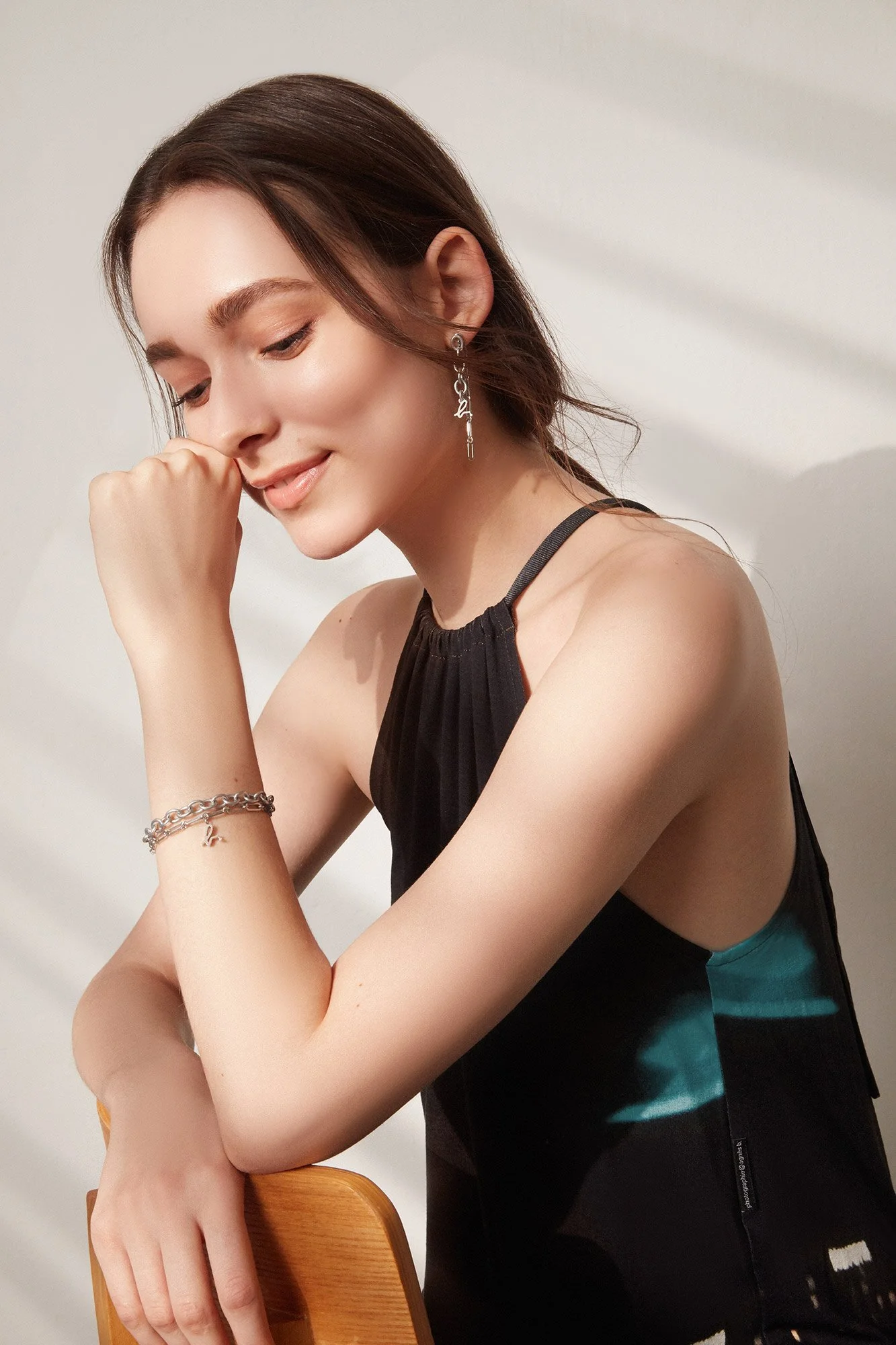 A young woman with dark brown hair and light skin, wearing earrings and a bracelet, is smiling with her eyes closed, resting her chin on her hand, in front of a plain light background.