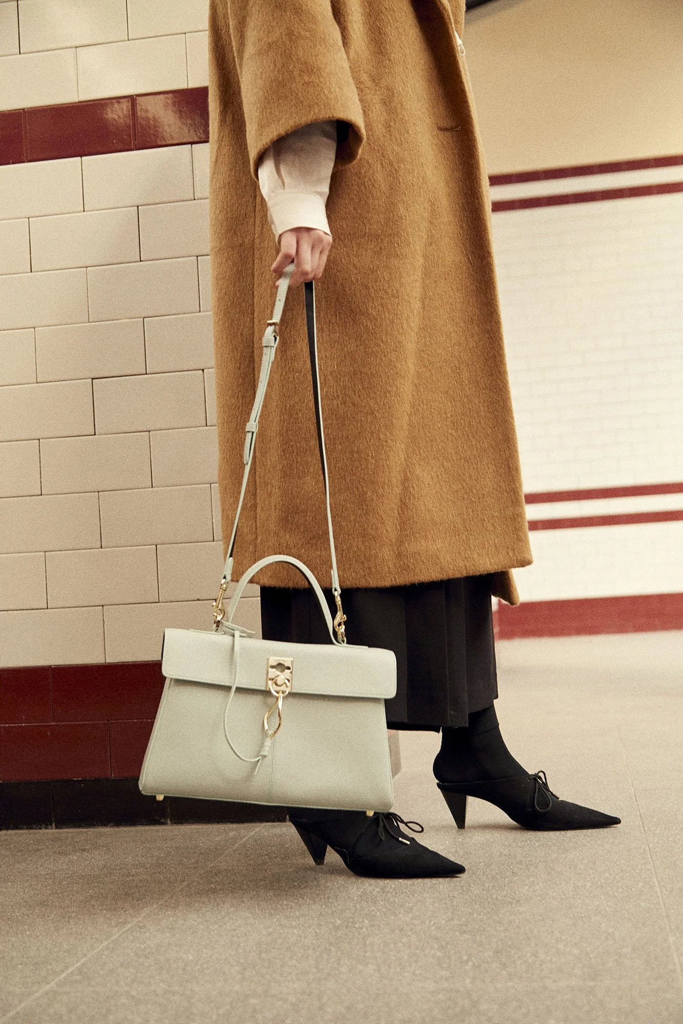 A woman in a brown coat and black pointed heels is standing indoors, holding a white handbag. The scene includes a tiled wall and a beige floor.