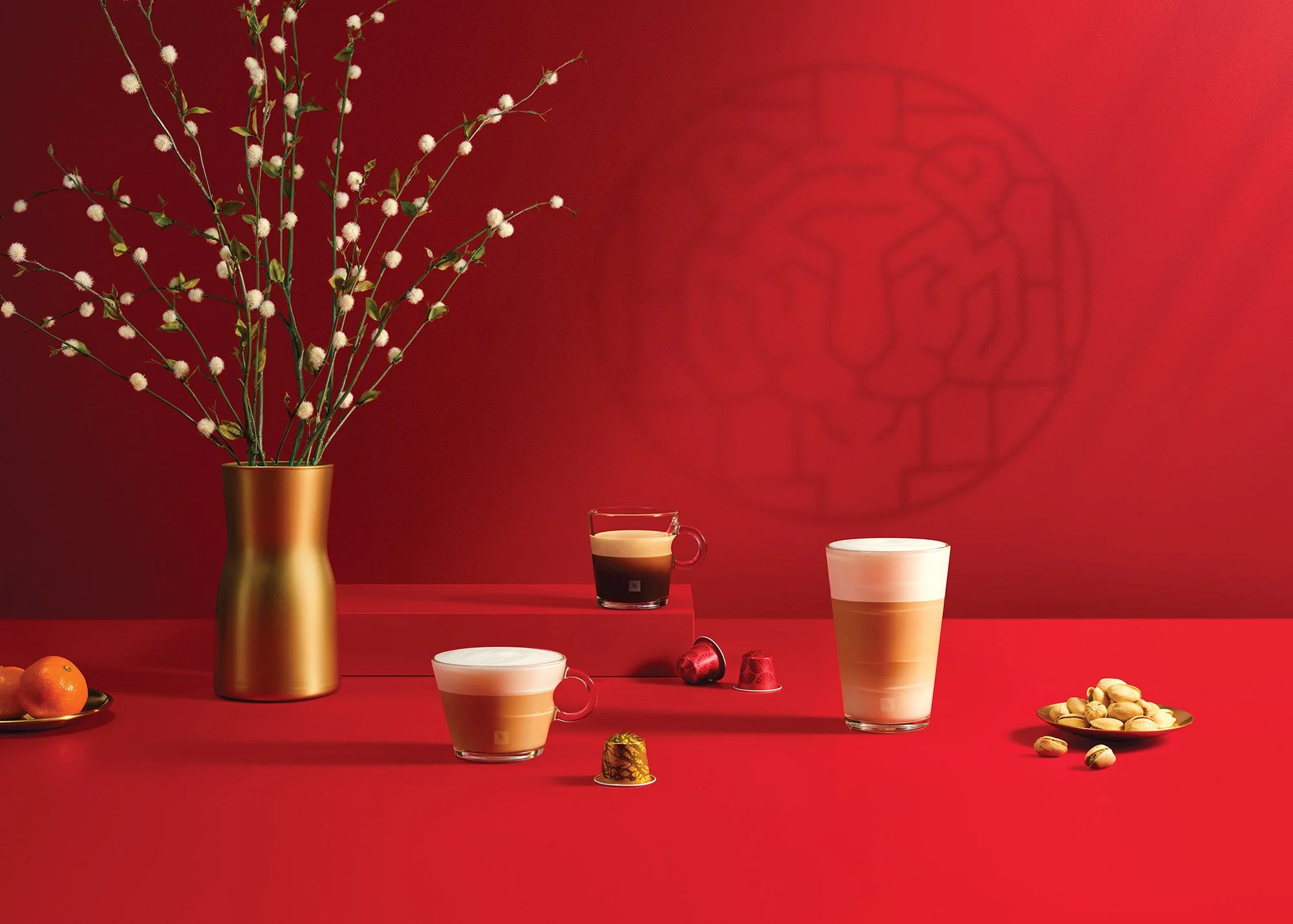 Red background with a gold vase holding tall white flowering branches, two cups of layered coffee drinks, a glass of dark coffee, a small plate of oranges, another plate of pistachios, and scattered coffee capsules.