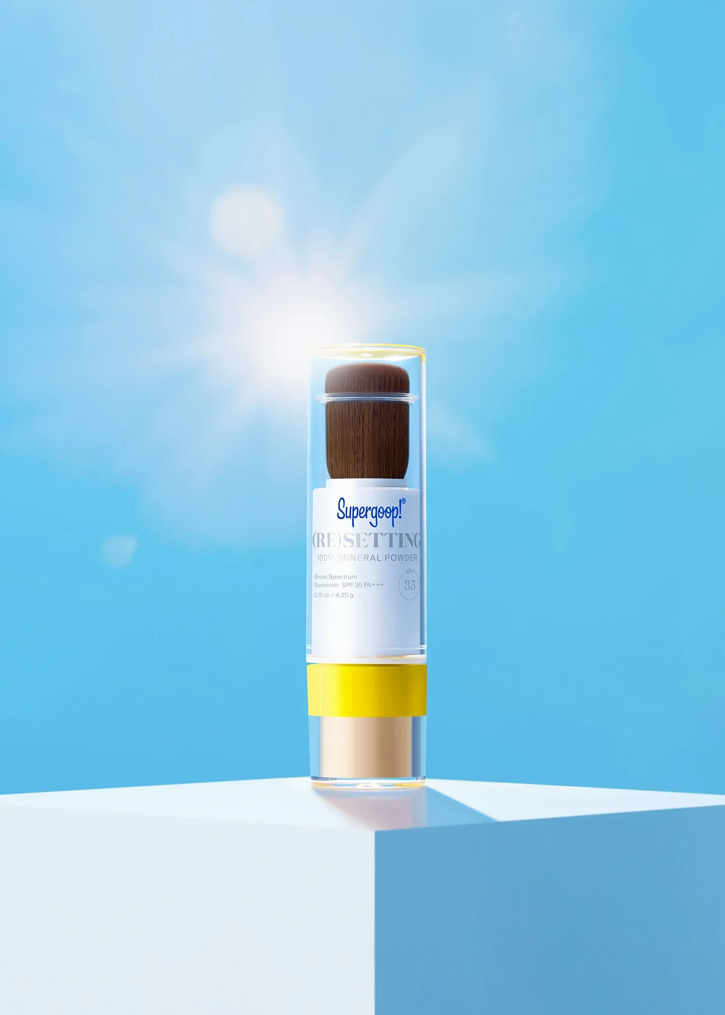 A bottle of Supergoop! (RE)SETTING mineral powder with a brush on top, placed on a white surface against a blue background with sunlight and lens flare.