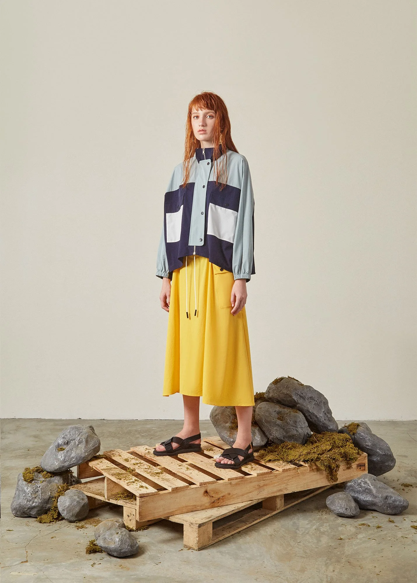 A woman standing on a wooden pallet platform surrounded by rocks and moss, wearing a color-blocked jacket with large pockets, yellow wide-leg pants, and black sandals.