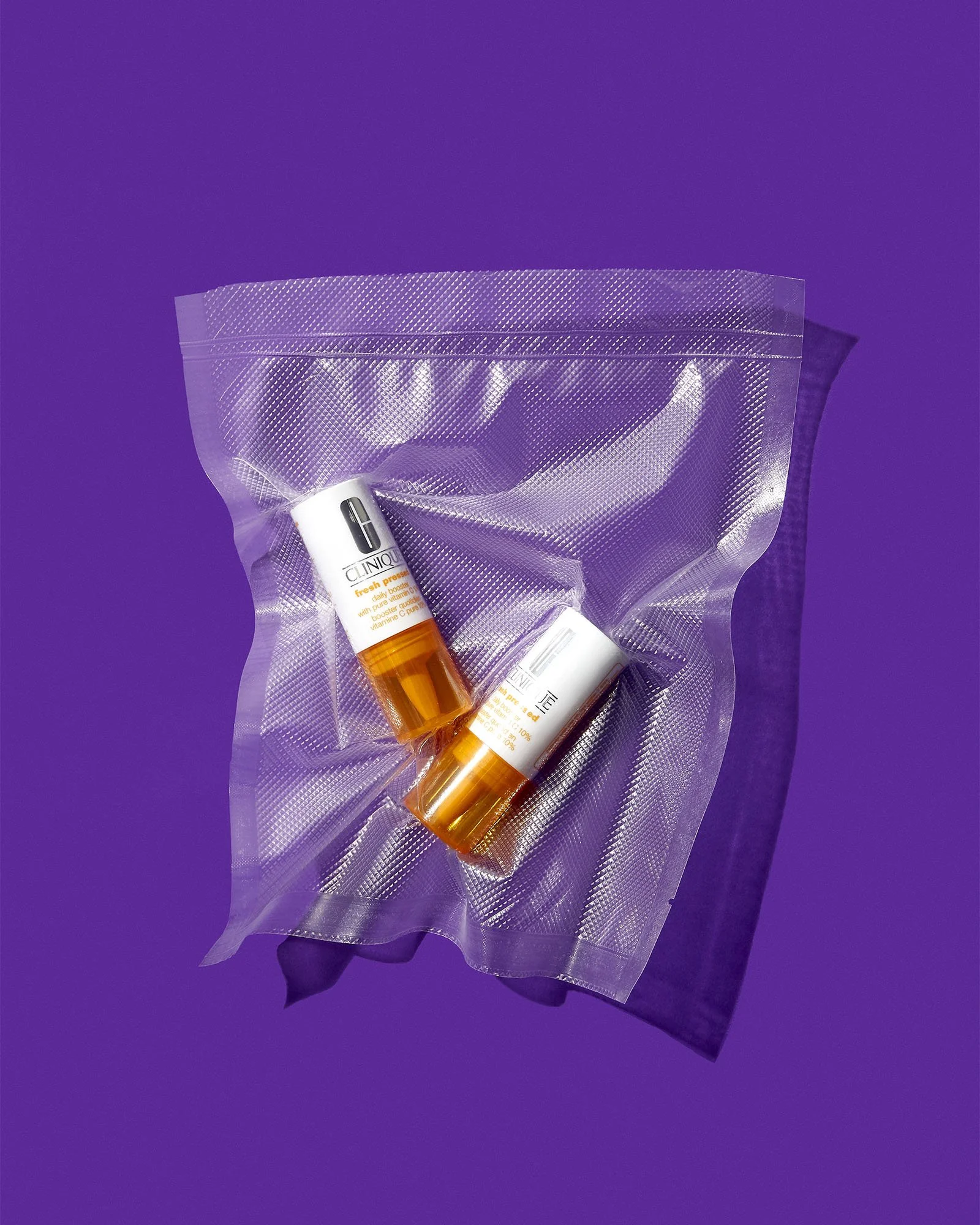 Two Clinique lip balm sticks inside a clear, resealable plastic pouch, placed on a purple surface with a black cloth partially visible underneath.