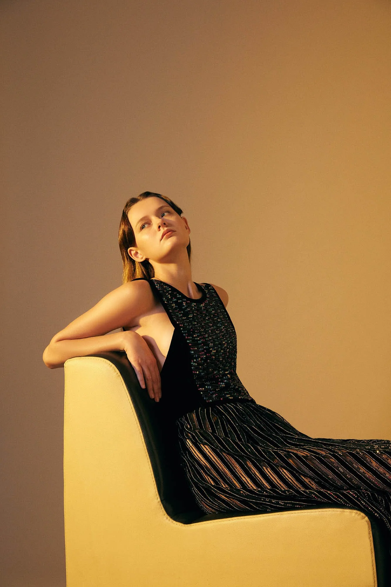 A young woman with brown hair styled in a vintage fashion, wearing a sleeveless black top with colorful embroidery and striped pants, sitting against a yellow chair with a neutral background, looking upward with a contemplative expression.