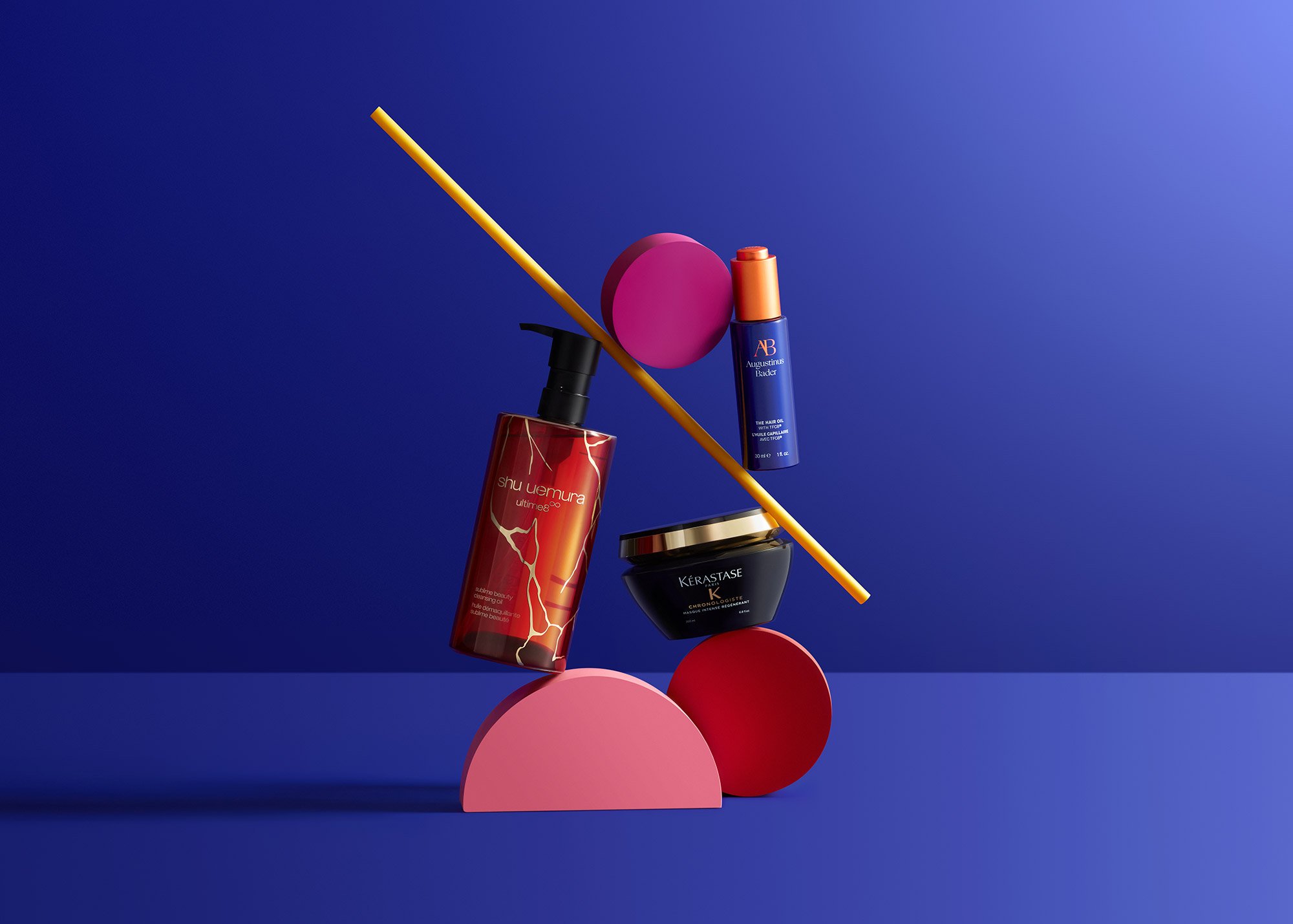 An artistic display of skincare and beauty products on colored geometric shapes against a blue background, including a red bottle with lightning design, a blue spray with orange cap, a black and gold jar, and a pink and purple sphere with a yellow st