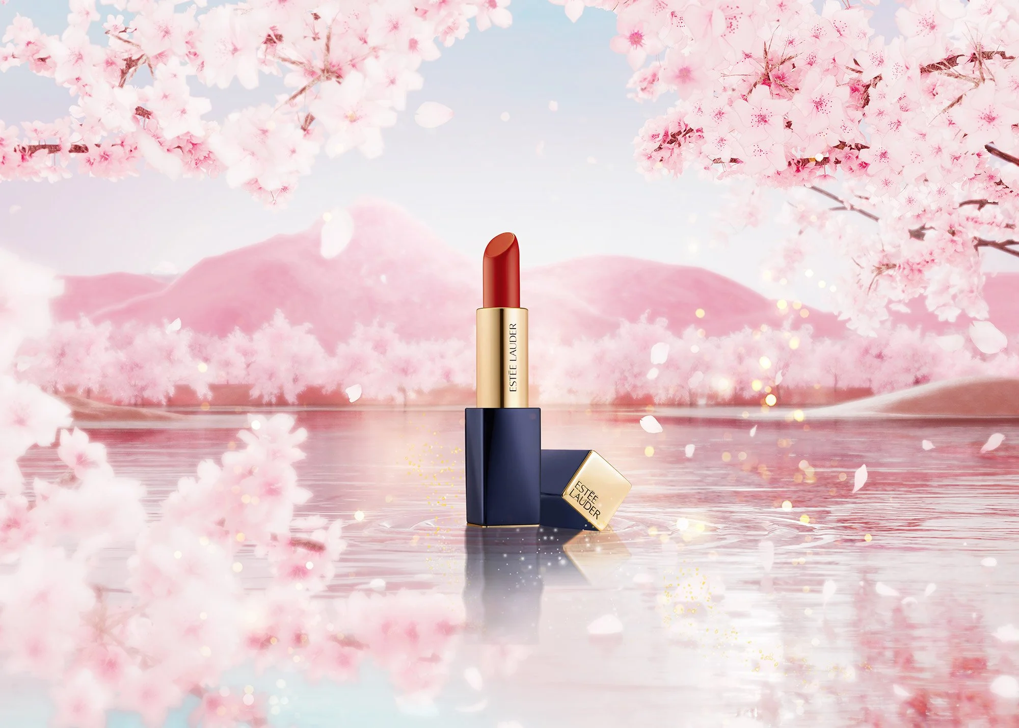 A lipstick with a gold and navy blue case, partially unscrewed to show red lipstick, is standing in a reflective water surface with pink cherry blossoms and mountain scenery in the background.