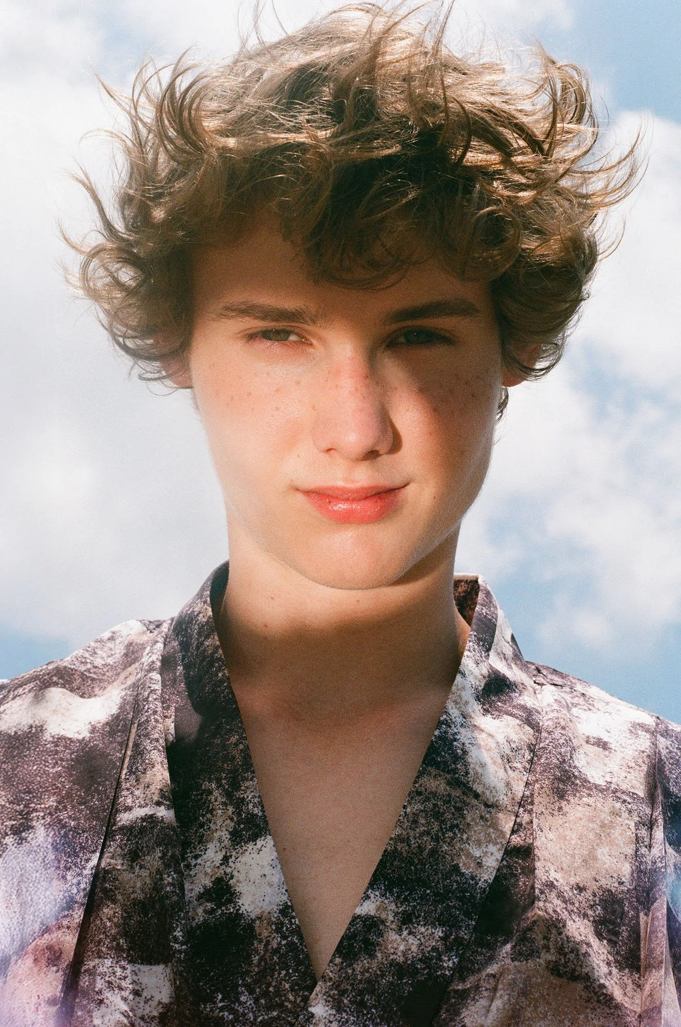 A young person with curly hair, freckles, and fair skin standing outdoors under a partly cloudy sky, wearing a patterned shirt.