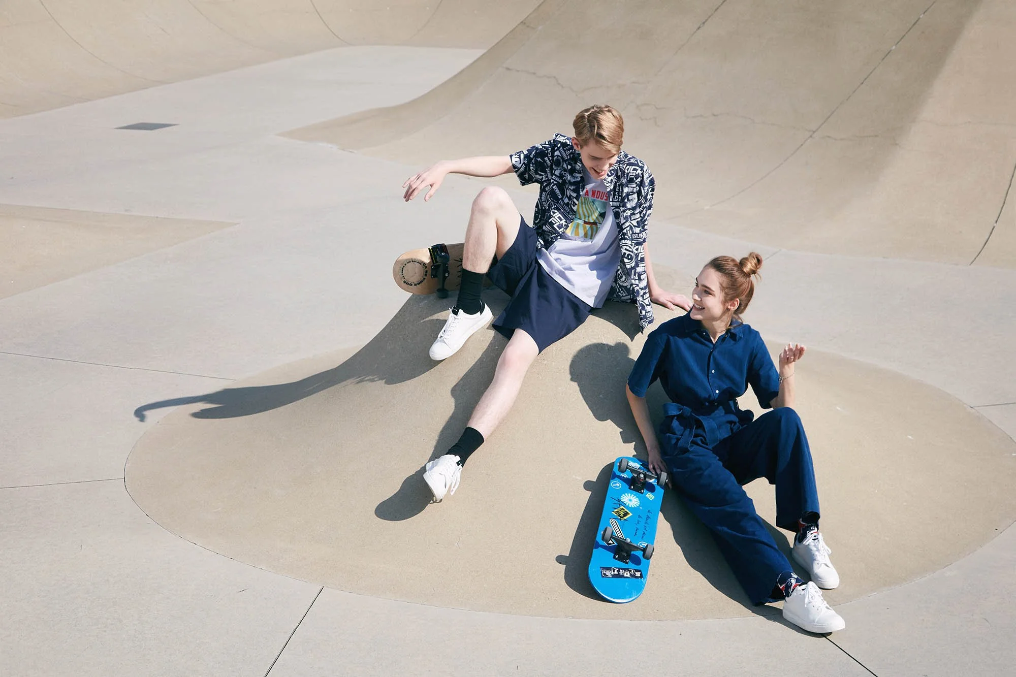 Two teenagers sitting on a skatepark bowl concrete surface, smiling and chatting with skateboards nearby.