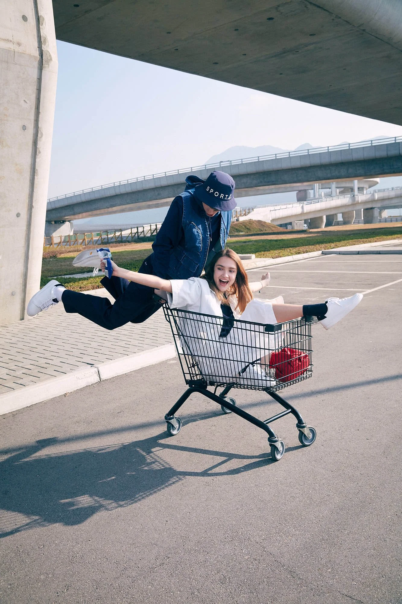 Two young women playing and having fun in a shopping cart outdoors near a highway overpass.