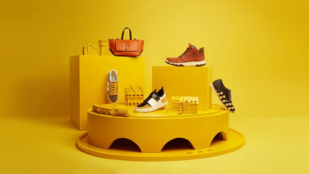 Display of colorful shoes and handbags on yellow platforms with yellow decorative buildings in the background.