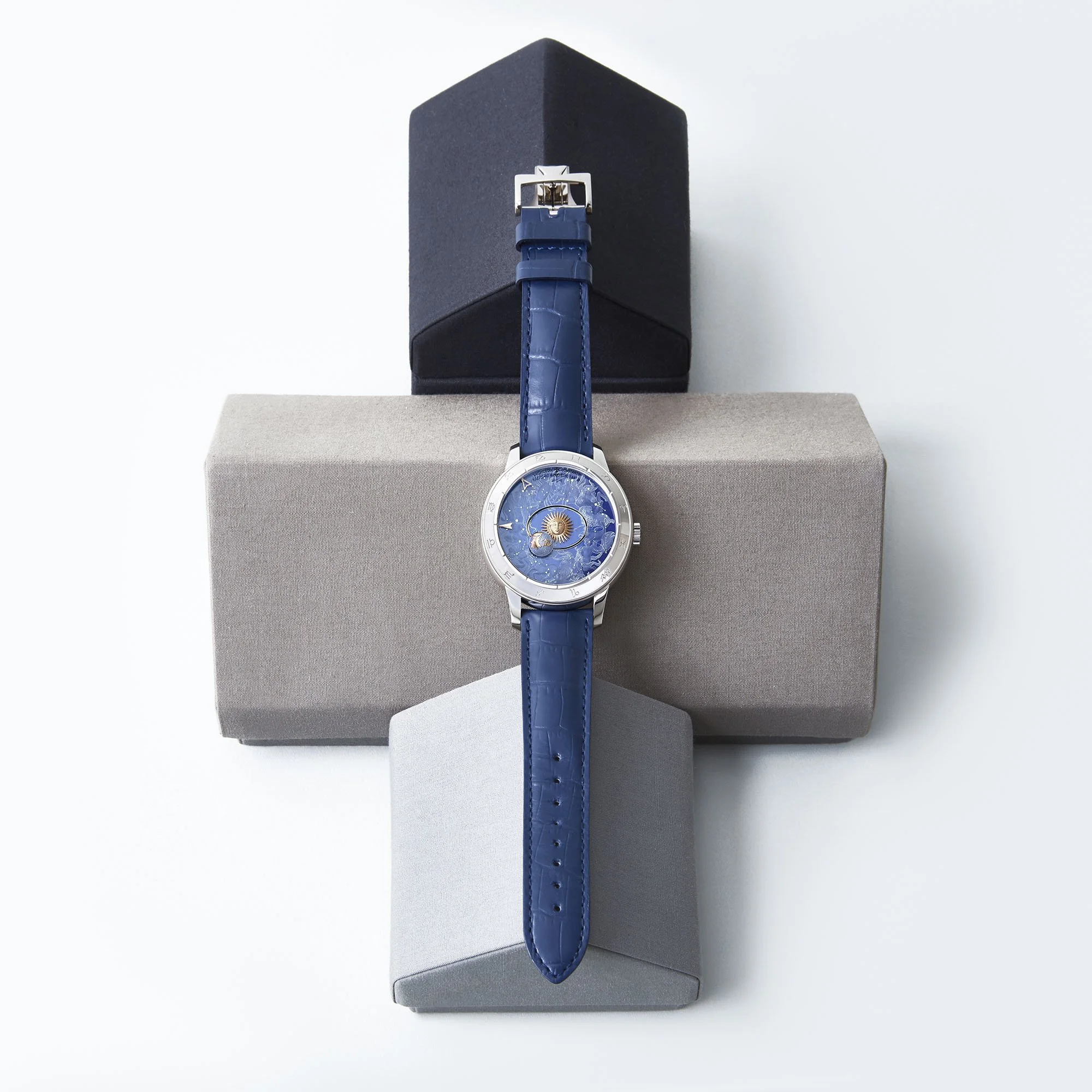 Luxury wristwatch with a blue strap and a detailed blue dial, displayed on a geometric stand with three stacked boxes in black, grey, and light grey background.