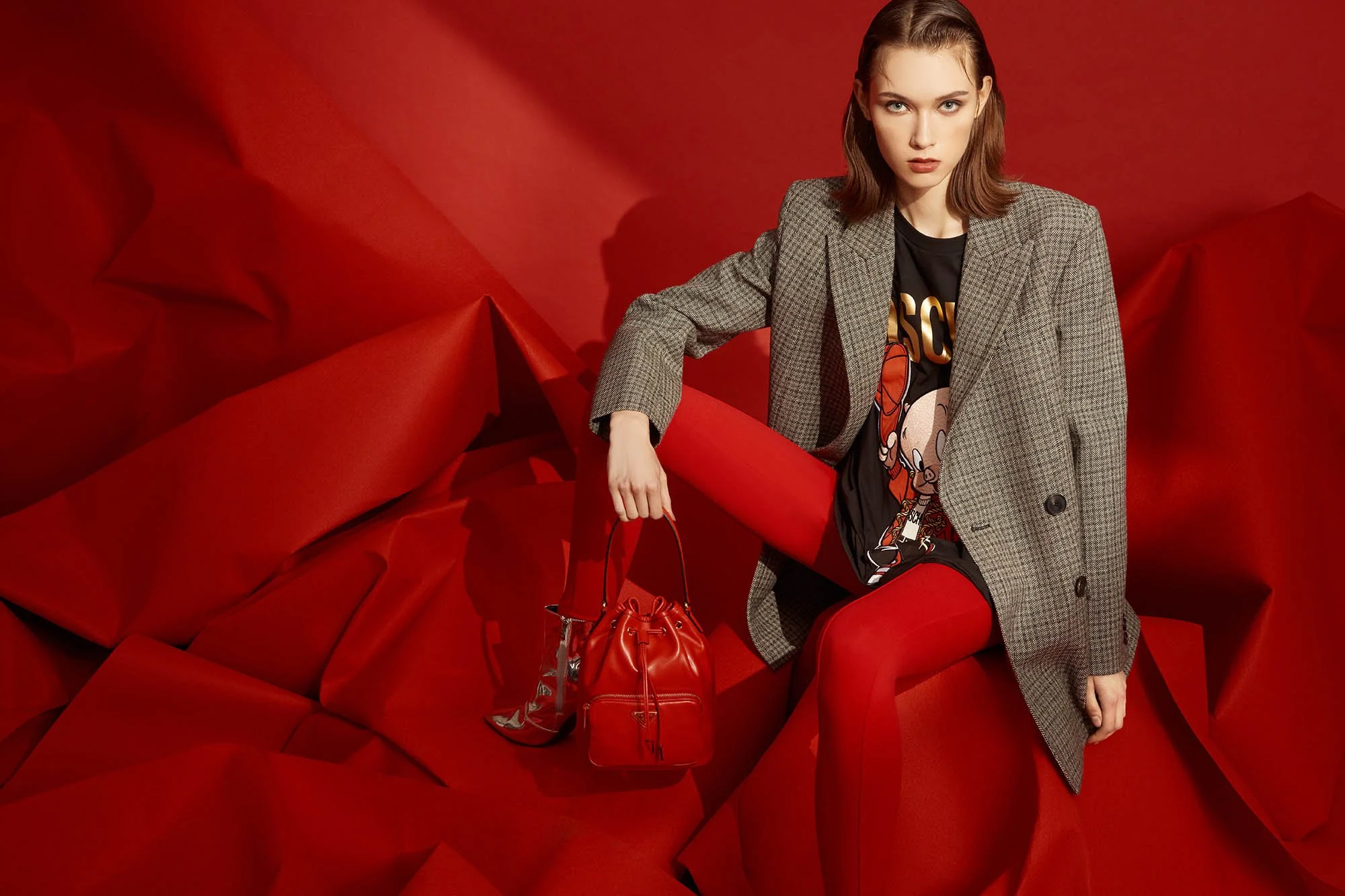 A woman with shoulder-length brown hair and blue eyes is sitting on a red, crumpled backdrop. She is wearing a gray plaid blazer over a black graphic t-shirt with cartoon characters, red tights, and red shoes. She is holding a small red handbag and h