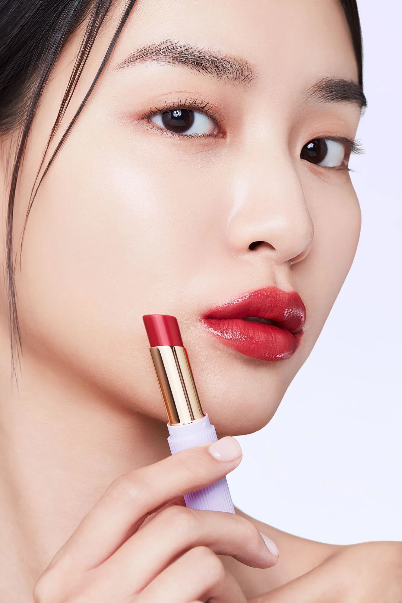 A close-up of a woman with smooth skin and dark hair, holding a lipstick close to her lips. She is wearing makeup, including red lipstick, eye makeup, and foundation.