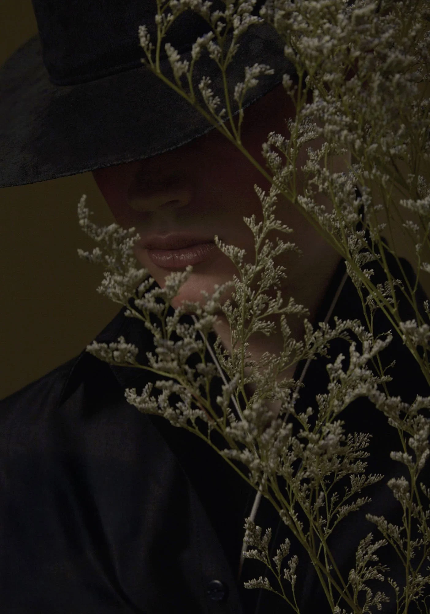 A woman wearing a large black hat with a bow, partially obscuring her face, surrounded by white flowering plants.