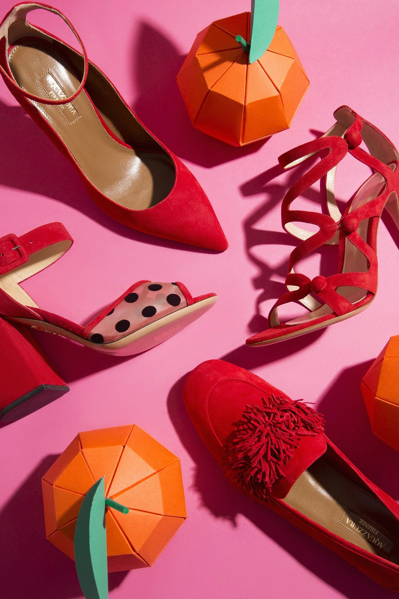 Red high heels, a pink and black polka dot shoe, red slingback heels, red dress shoe with fringe detail, and two orange and green paper umbrellas on a pink background.