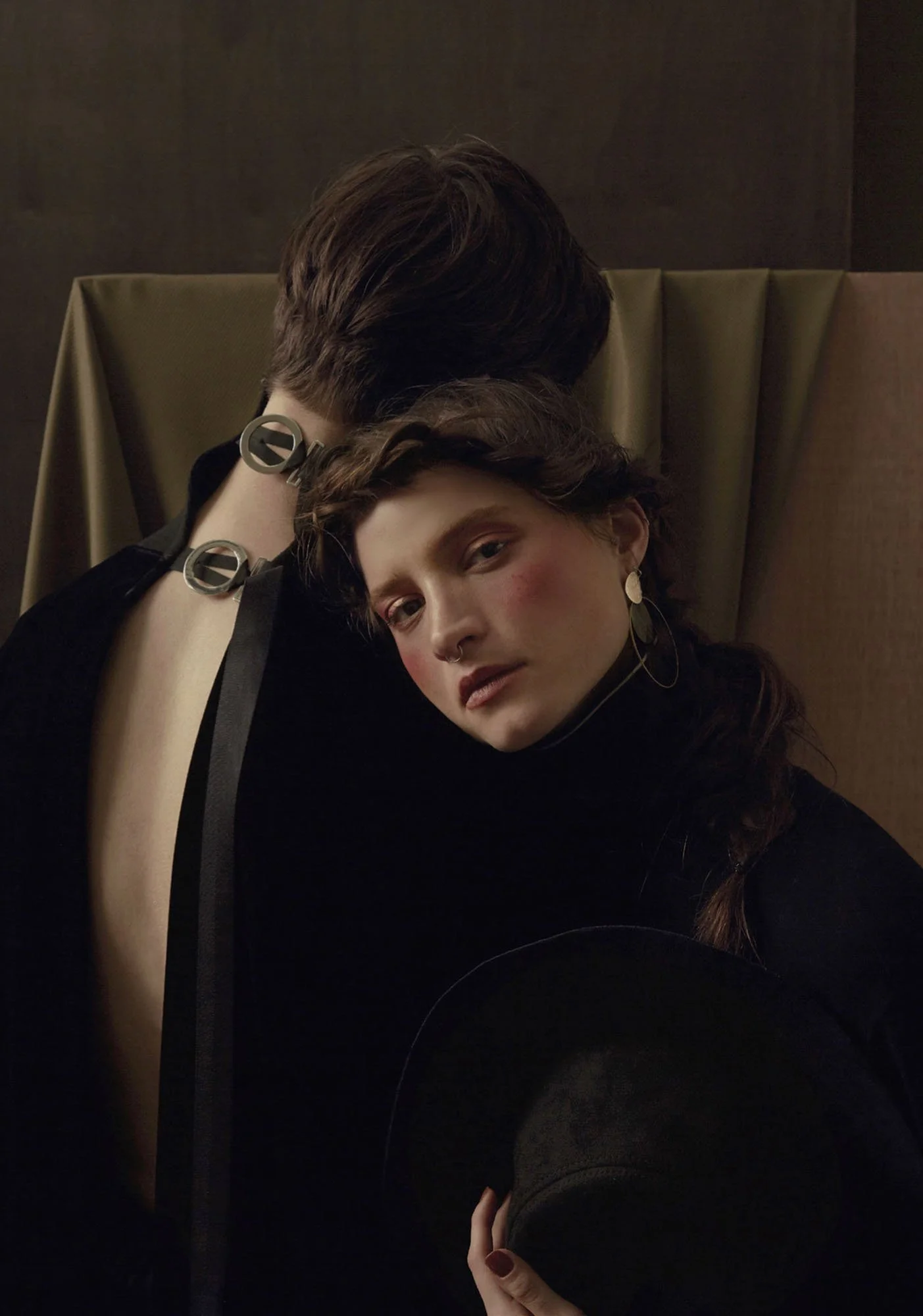 A woman with earrings and a nose ring, lying on a tan lounge, resting her head on a person with short hair, both dressed in black, with a dark background.