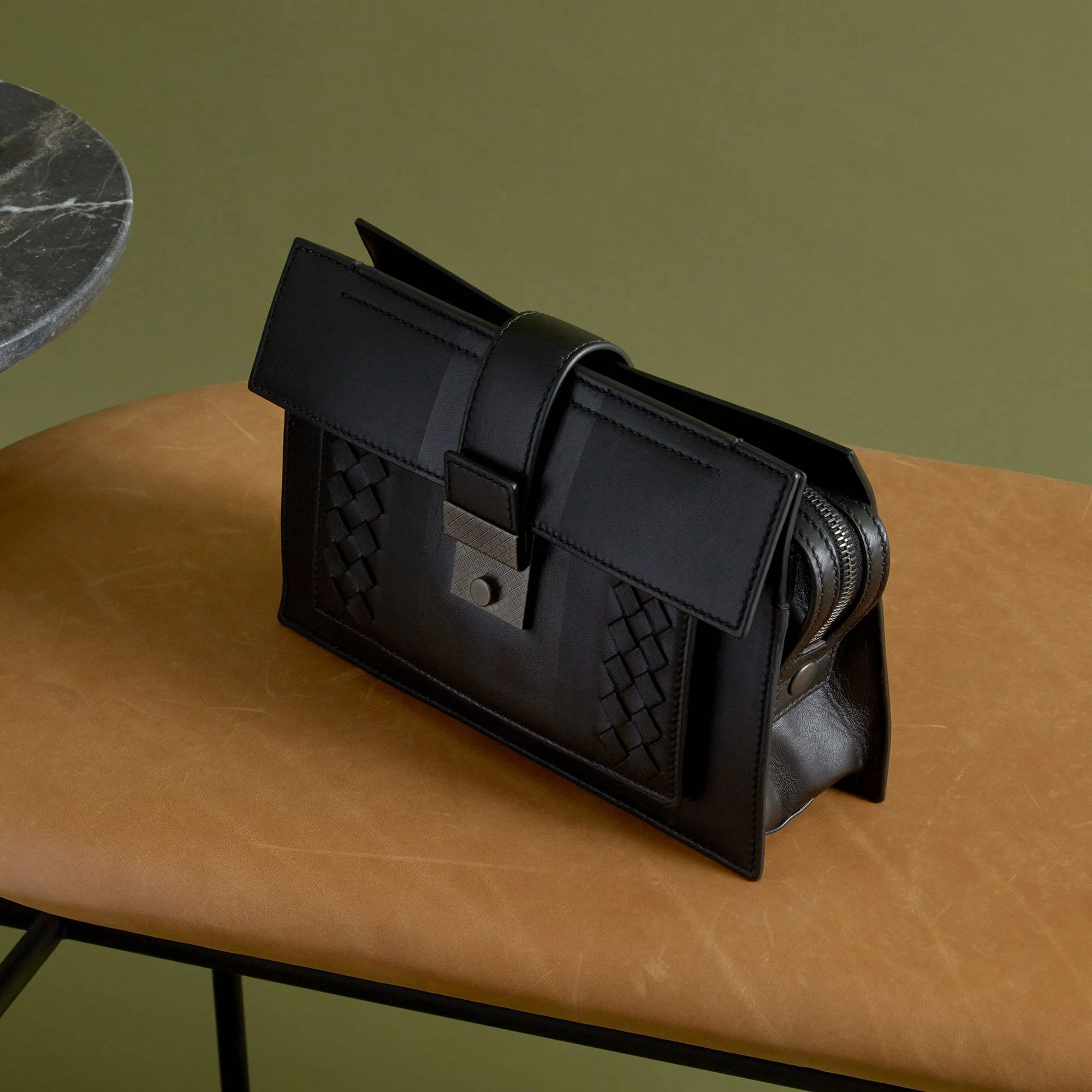 Black leather handbag with woven details and a silver clasp, placed on a brown leather bench with a green wall in the background.