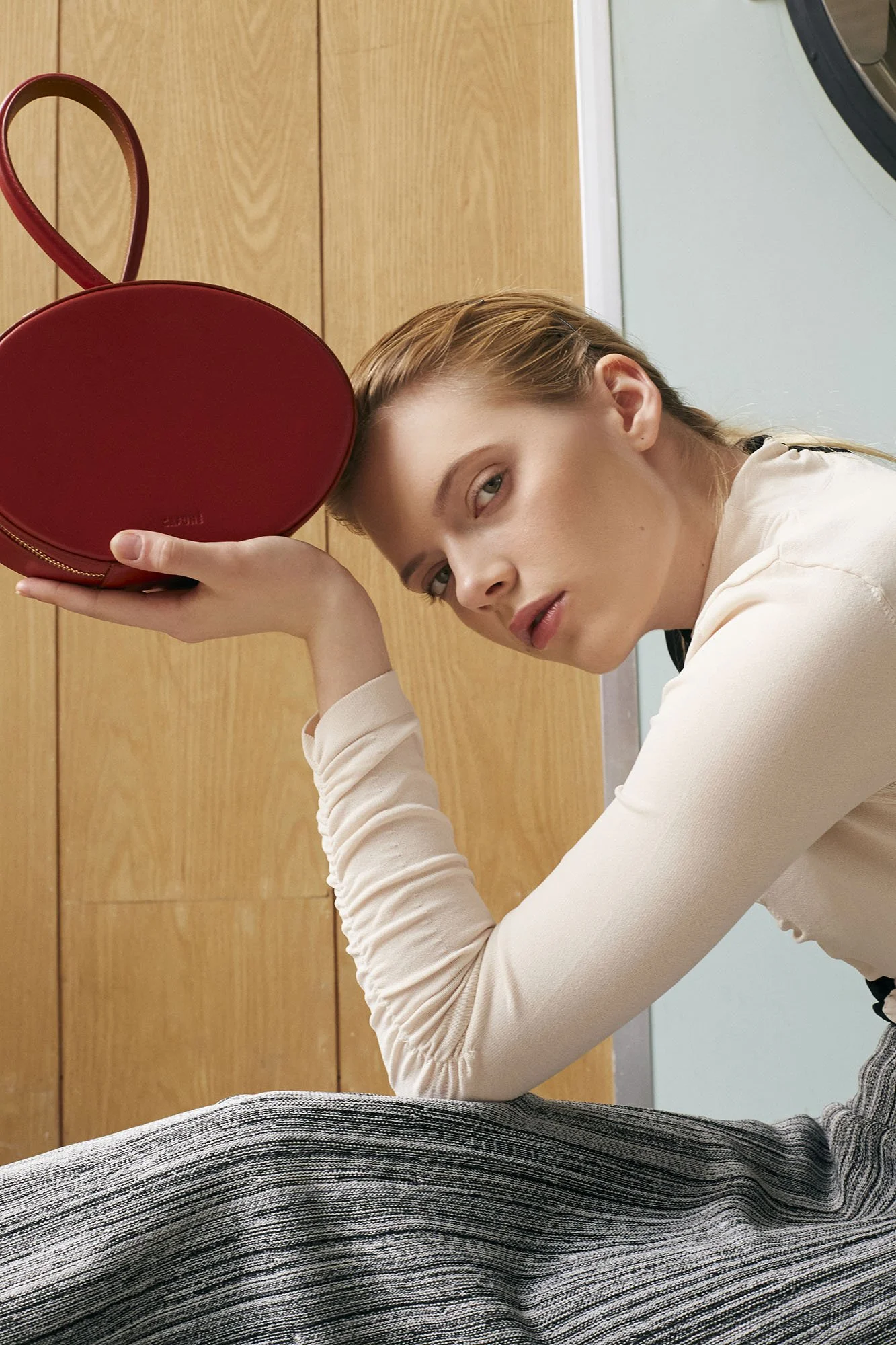 A young woman with blonde hair and blue eyes, wearing a beige long sleeve top, resting her head on a red circular object and looking at the camera, in a room with blue wall and wooden cabinets.