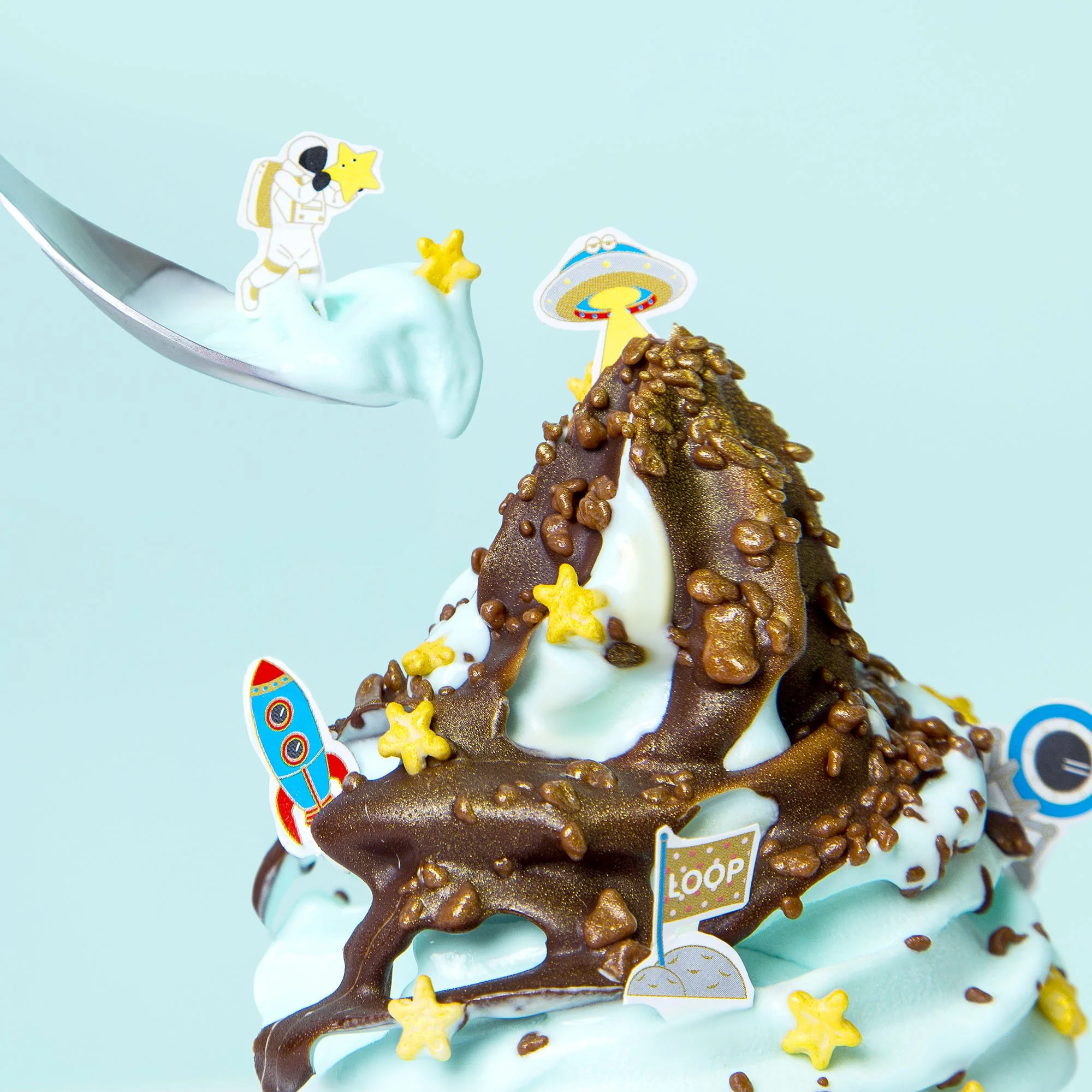 Close-up of a pastel blue frosted cake decorated with chocolate, yellow stars, and space-themed toppers including an astronaut, planets, a UFO, and a rocket.