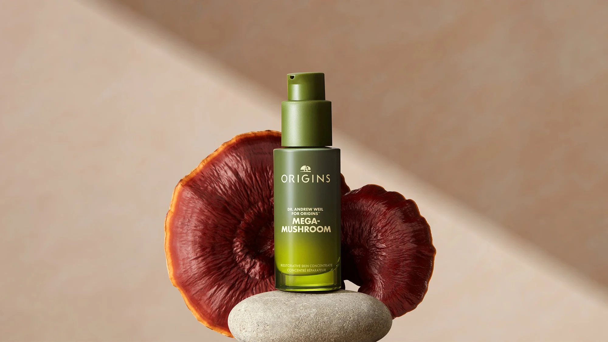 A green bottle of Origins Mega Mushroom skincare product resting on a gray stone, with a dried red mushroom behind it, on a beige background.