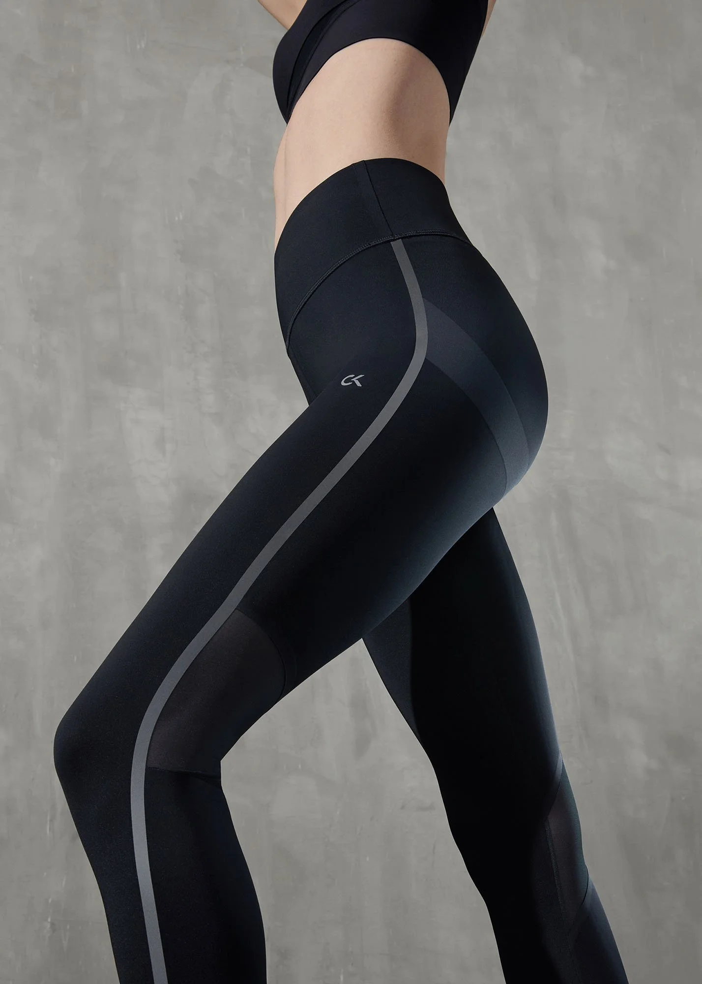 Close-up of a person in black workout leggings with gray accents, standing with one leg crossed over the other against a concrete background.