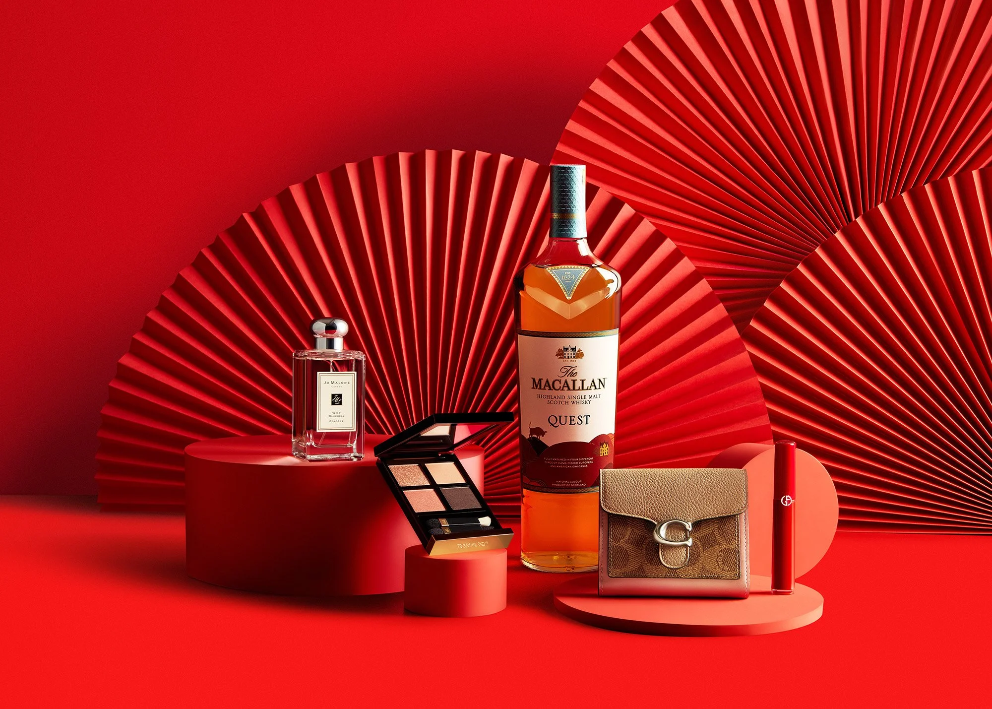 Red background with large red fans, displaying a bottle of Macallan Scotch whisky, a Jo Malone perfume, a Tom Ford eyeshadow palette, a small designer wallet, and a red lipstick.