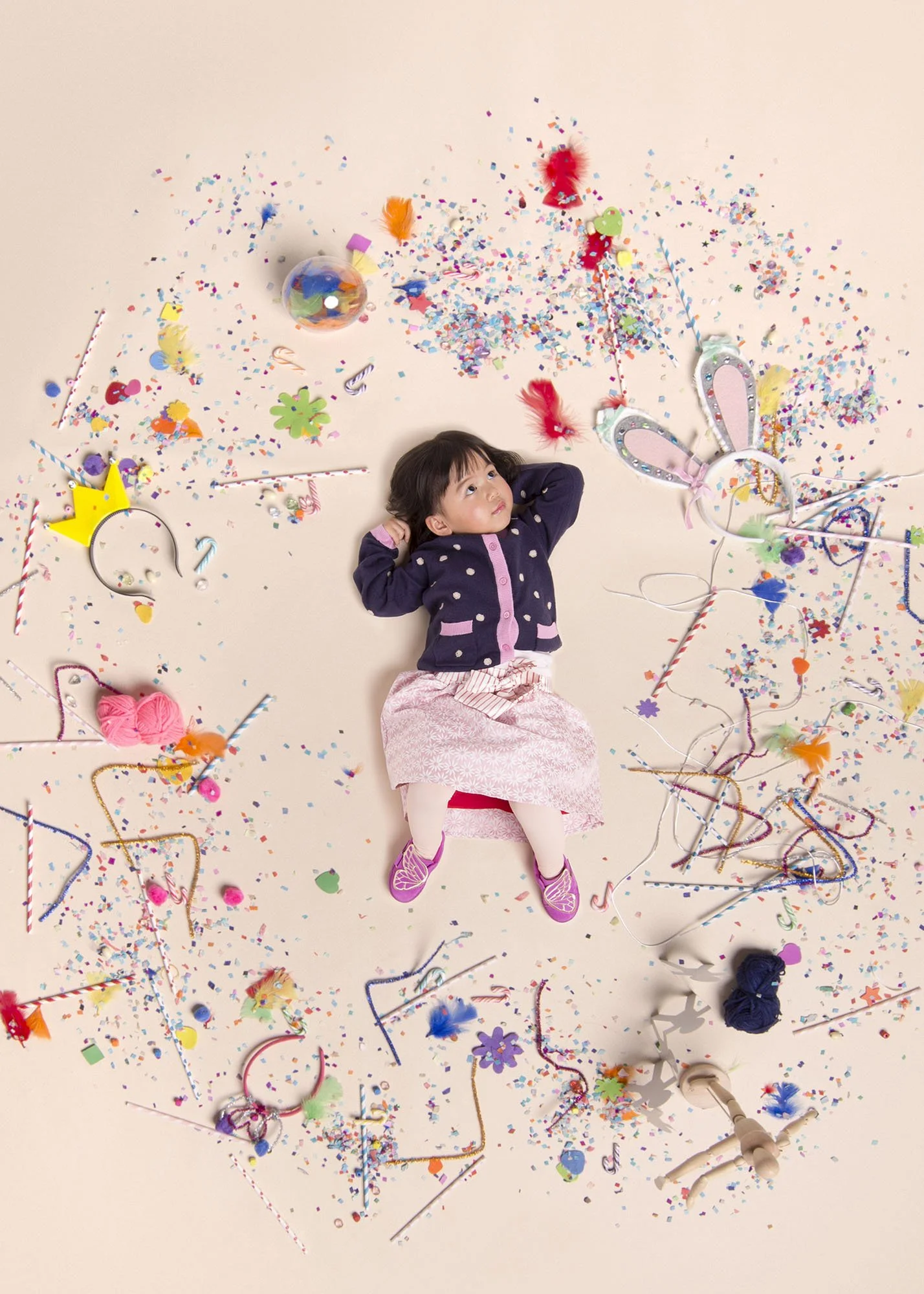 A young girl lying on a beige background surrounded by colorful confetti, party streamers, and decorations like balloons and paper cutouts, looking up with her hands behind her head.