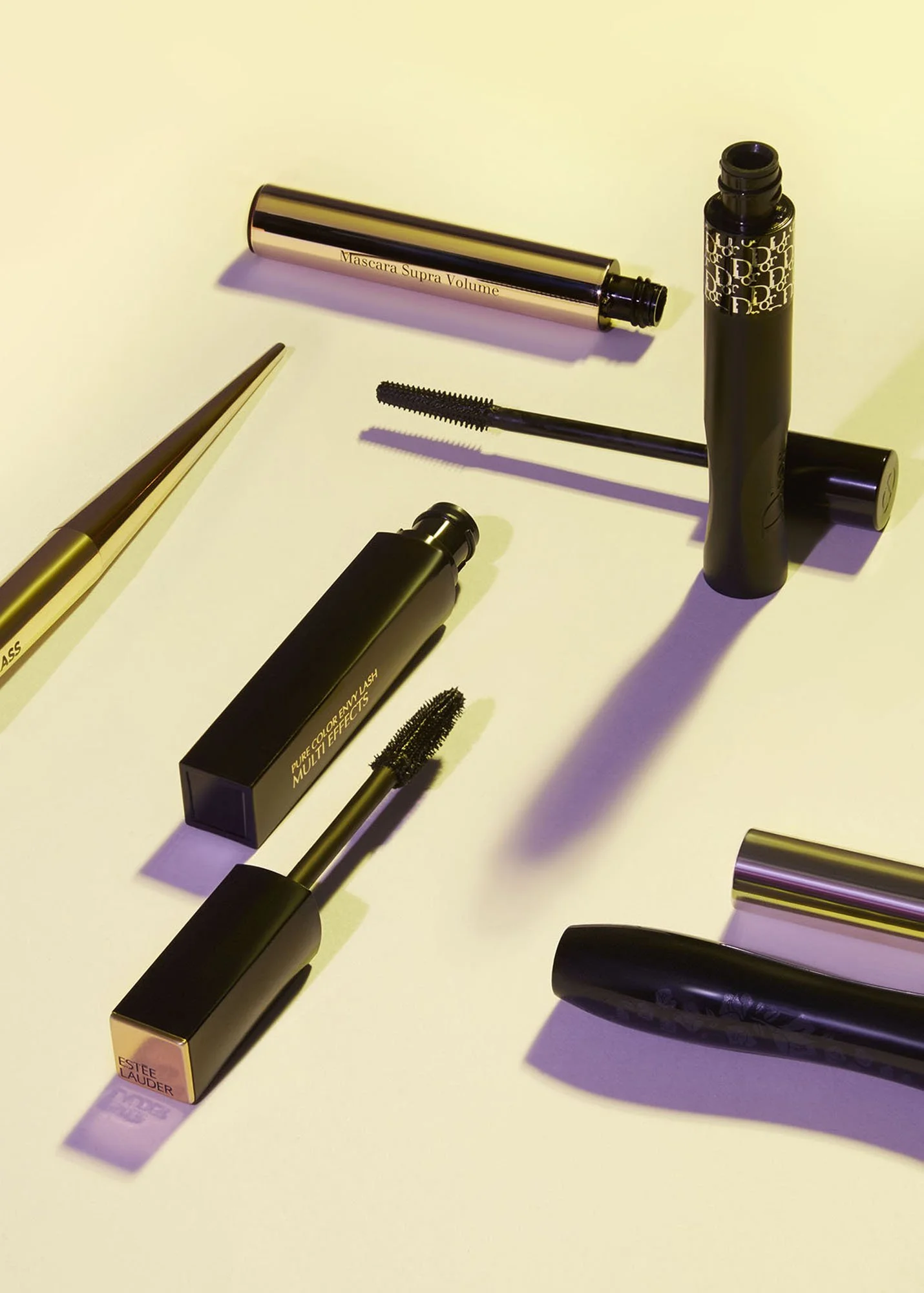 Collection of makeup products including mascara, lipstick, and eye liner, arranged on a white surface with shadows cast in a warm light.