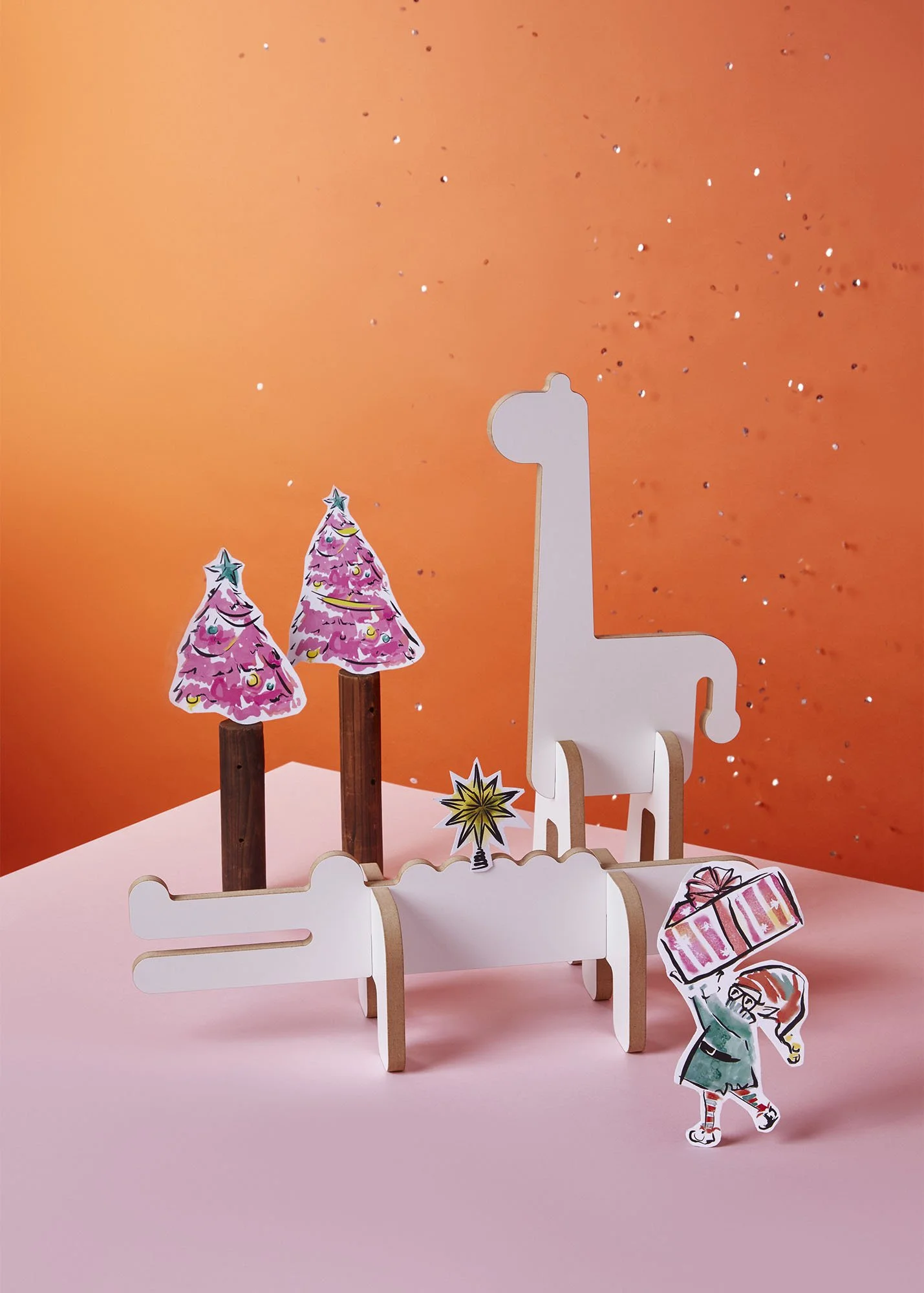 Decorative Christmas scene with cardboard cutouts of pink Christmas trees, a white giraffe-shaped display, a cartoon girl carrying a wrapped gift, and a gold star on a pink and orange background with glitter.