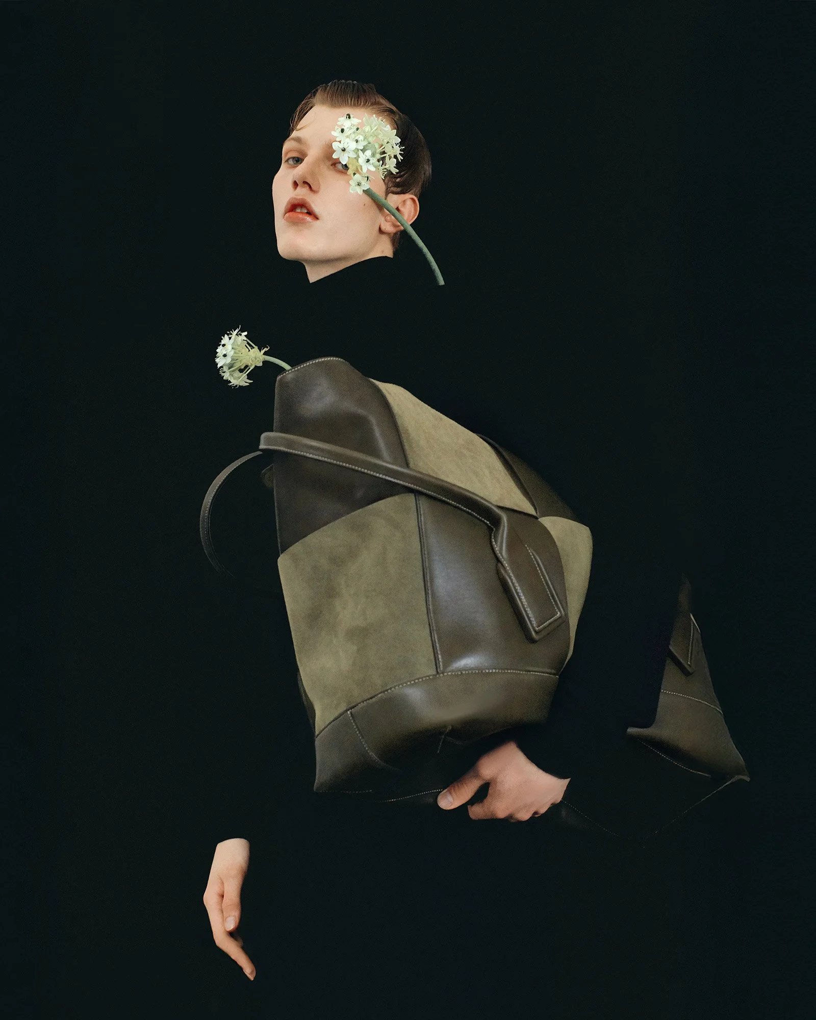 A person with short, styled hair holding a large two-tone brown and khaki bag with black clothing, with two white flowers near their face against a dark background.