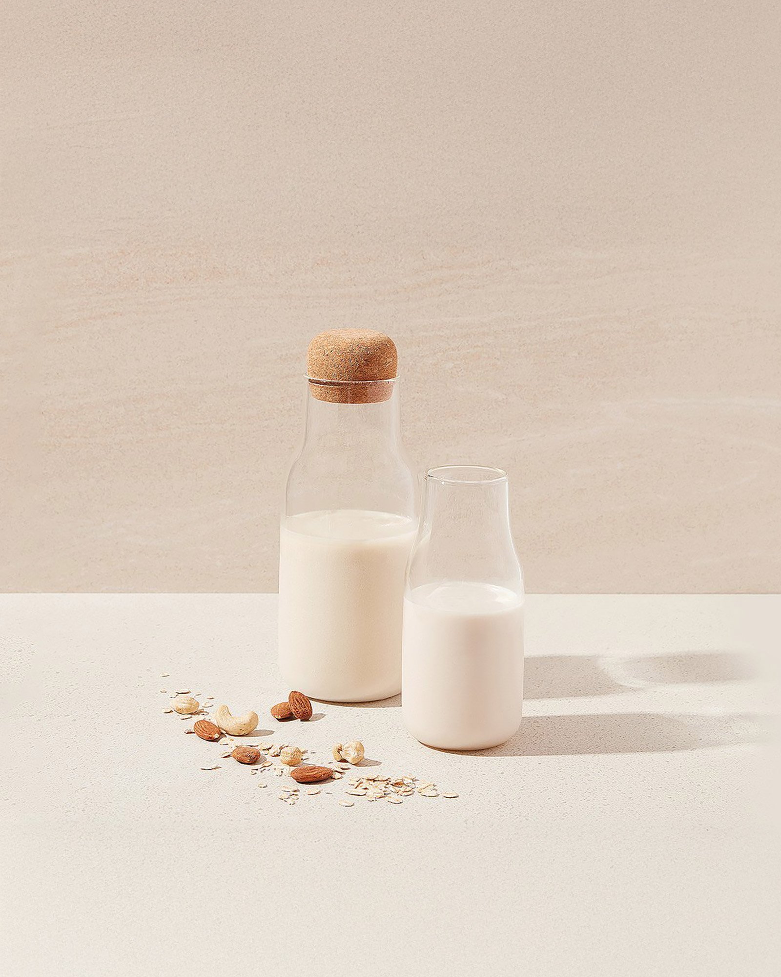 A glass bottle and a glass filled with milk, with scattered almonds and oats on a white surface in a minimalistic setting.