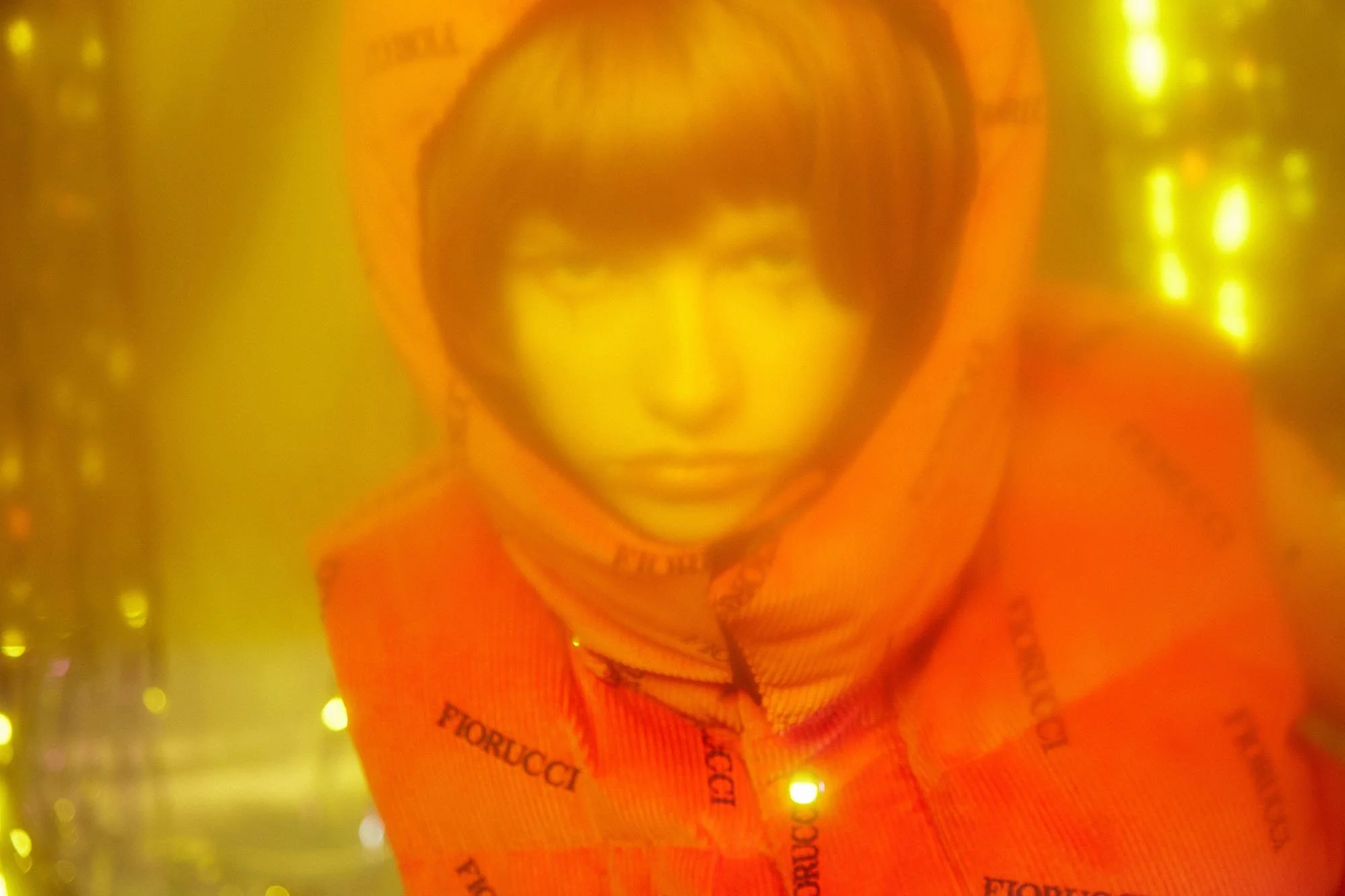 A person with short hair wearing an orange hoodie with the word 'Fiorucci' printed repeatedly on it, surrounded by yellow lights creating a warm, hazy effect.
