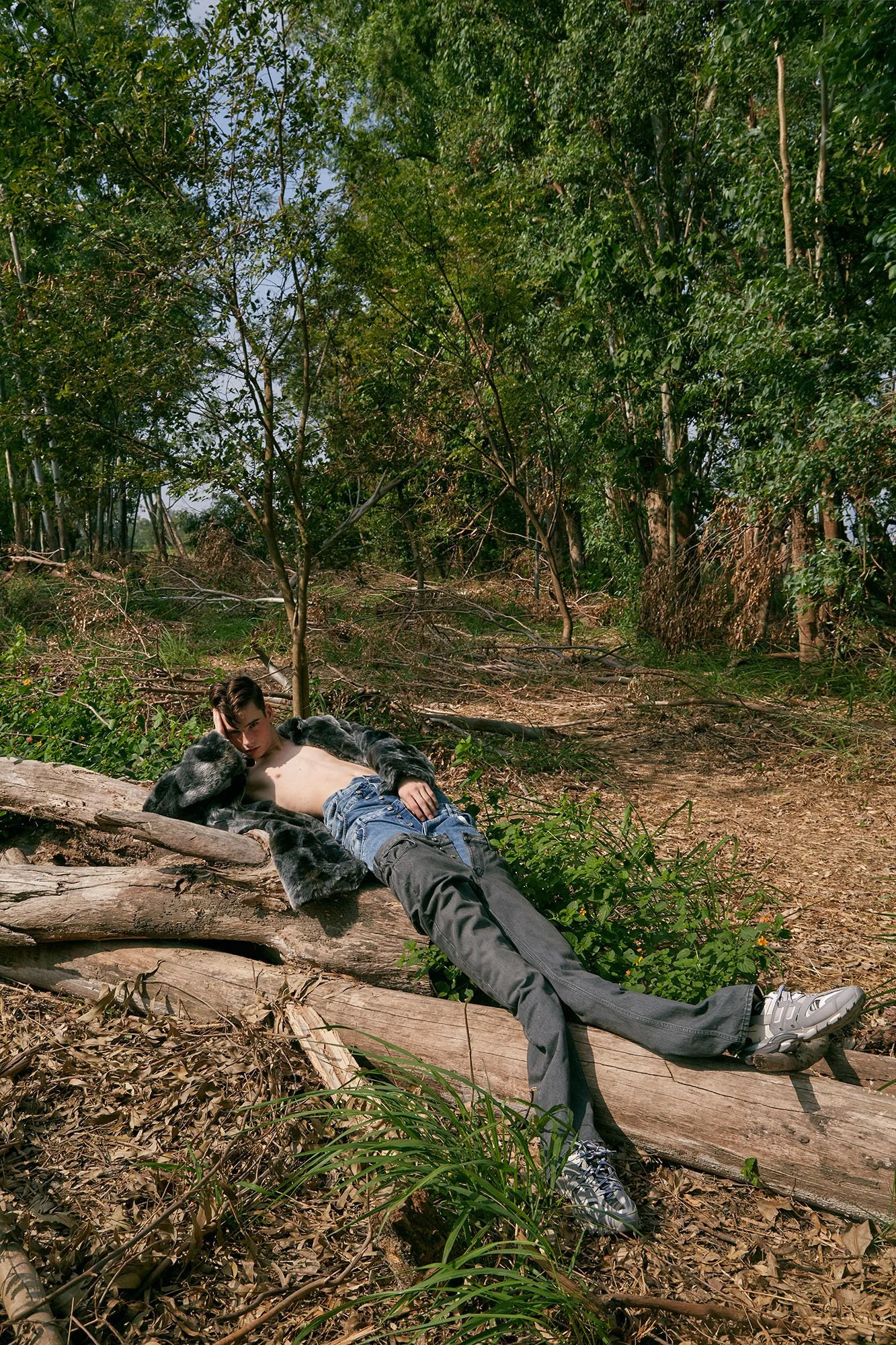 Man lying shirtless on a fallen log in a forest, wearing black pants and white sneakers, with lush greenery and trees in the background.