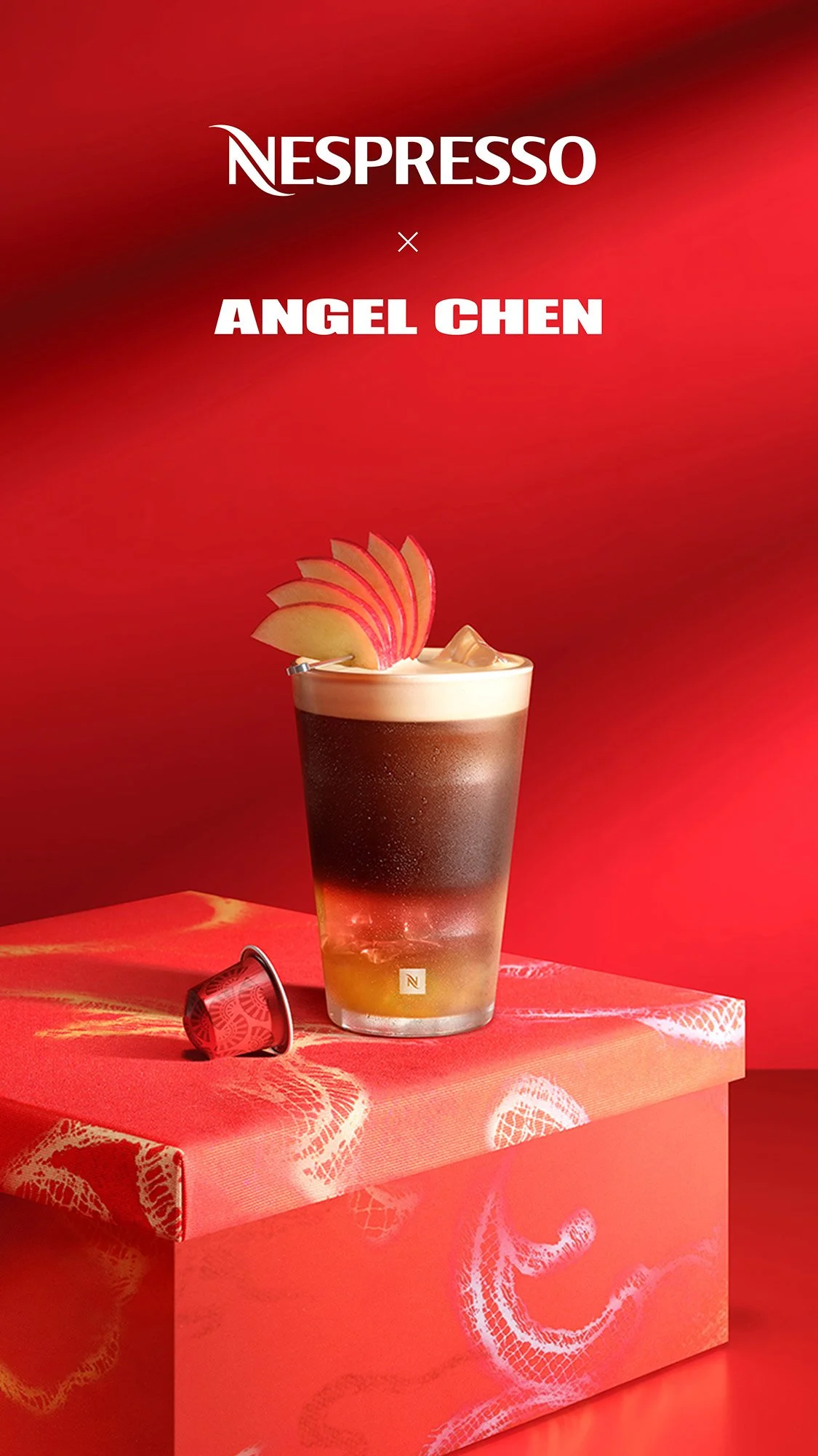 A promotional image for Nespresso featuring Angel Chen. The image shows a layered coffee drink with foam, garnished with apple slices, placed on a red box with white decorative patterns. The background is red with soft gradients.