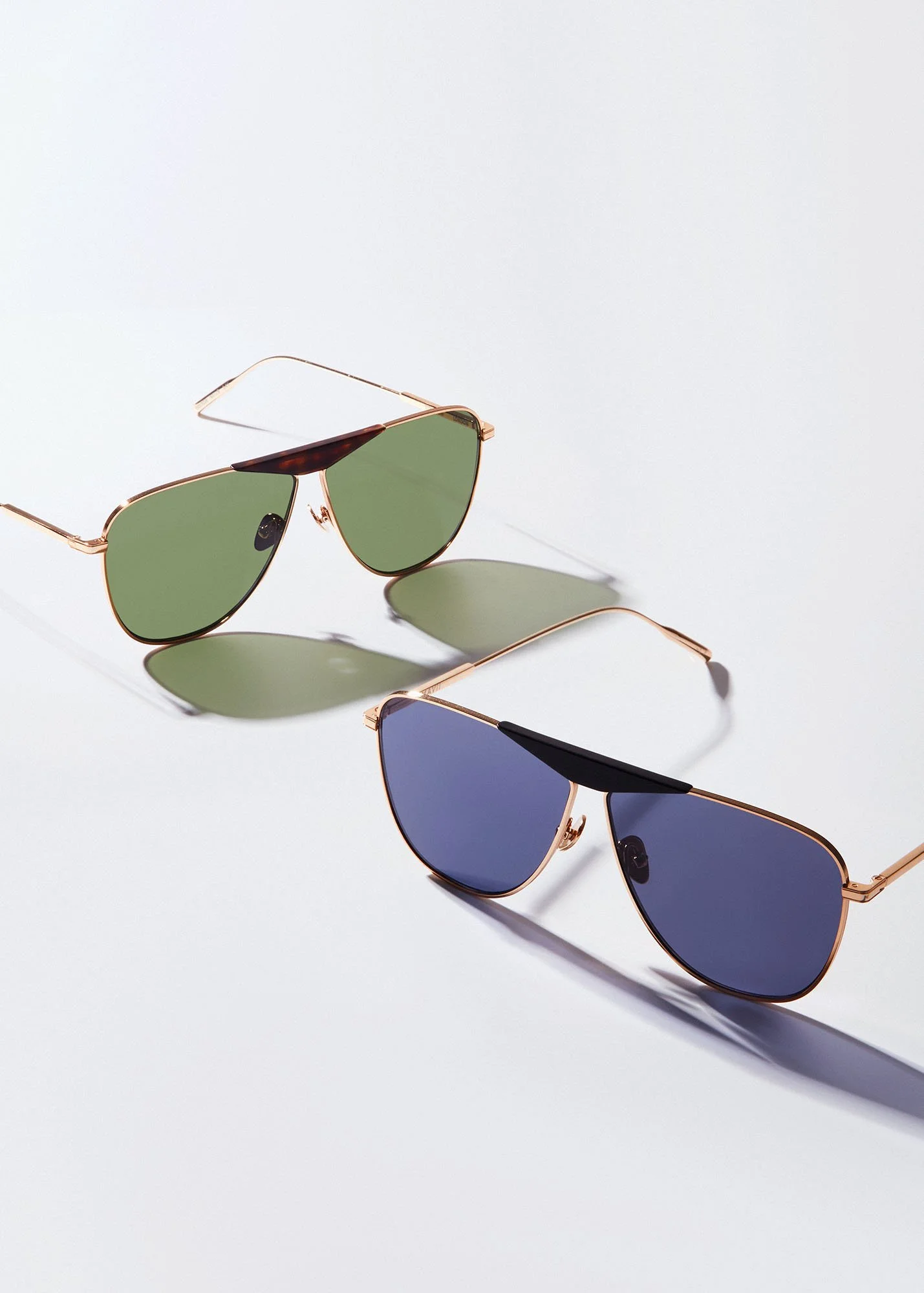 Three pairs of sunglasses with different colored lenses and metal frames are arranged on a white surface, casting shadows.