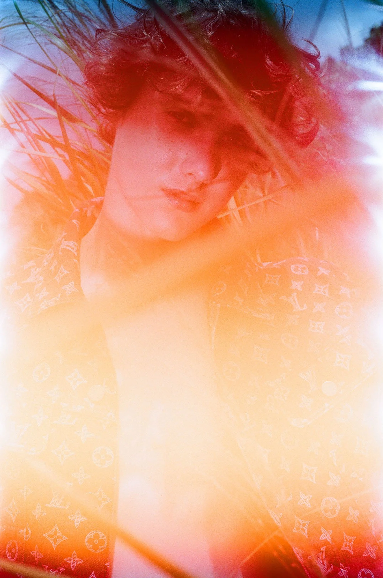 A person with curly hair lying in tall grass, wearing a Louis Vuitton patterned jacket, with sunlight creating a colorful, dreamy effect.