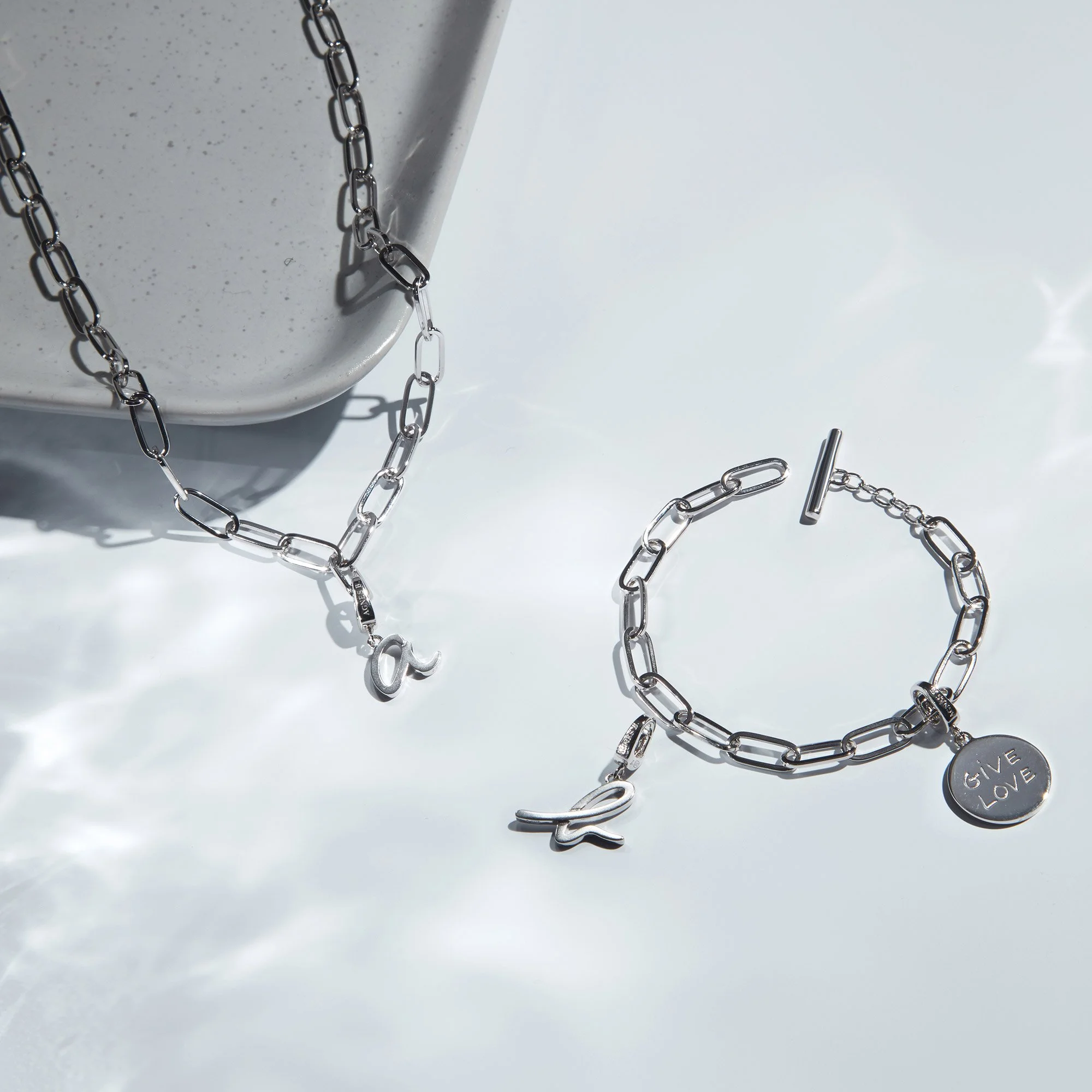Silver jewelry set consisting of a chain necklace with a charm, a bracelet with a charm that says 'GIVE LOVE', and a second bracelet with a small charm, all placed on a reflective surface near a speckled gray dish.