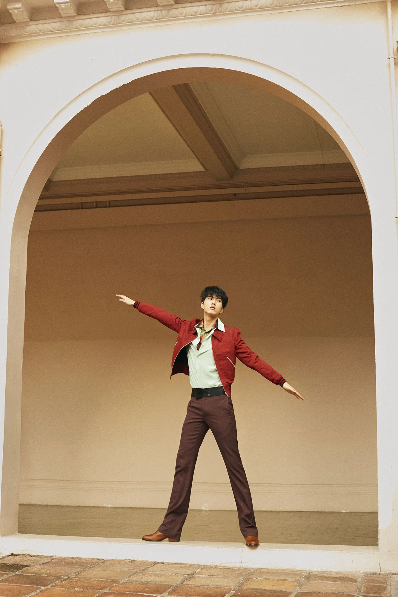 A young man standing in a large archway, striking a dance or pose with arms extended to the sides, wearing a red jacket, light-colored shirt, dark pants, and brown shoes, against a plain beige background.