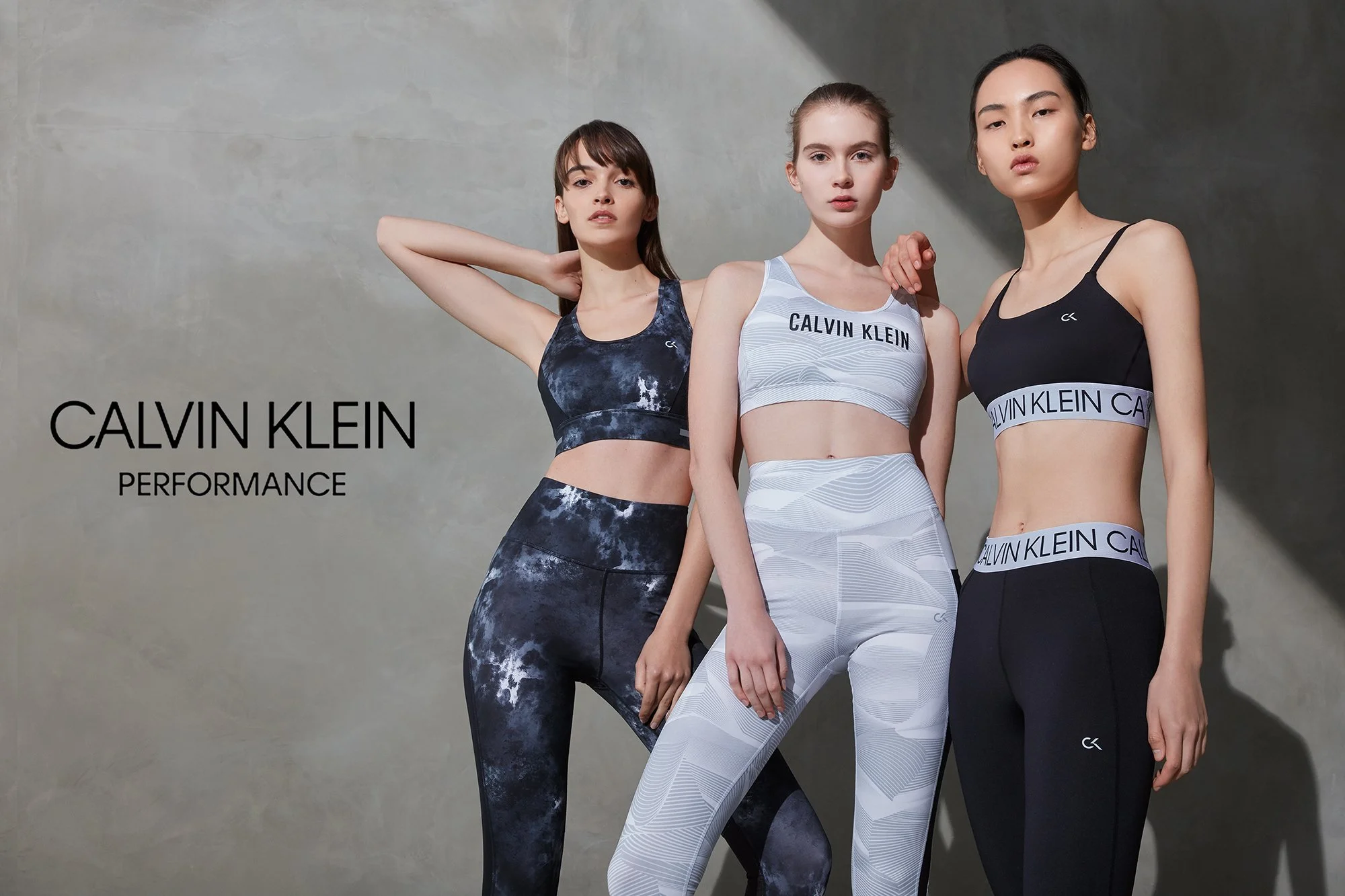Three women modeling Calvin Klein sportswear. The woman on the left wears a blue tie-dye sports bra and matching leggings. The woman in the middle wears a light gray sports bra and matching leggings with wave patterns. The woman on the right wears a 