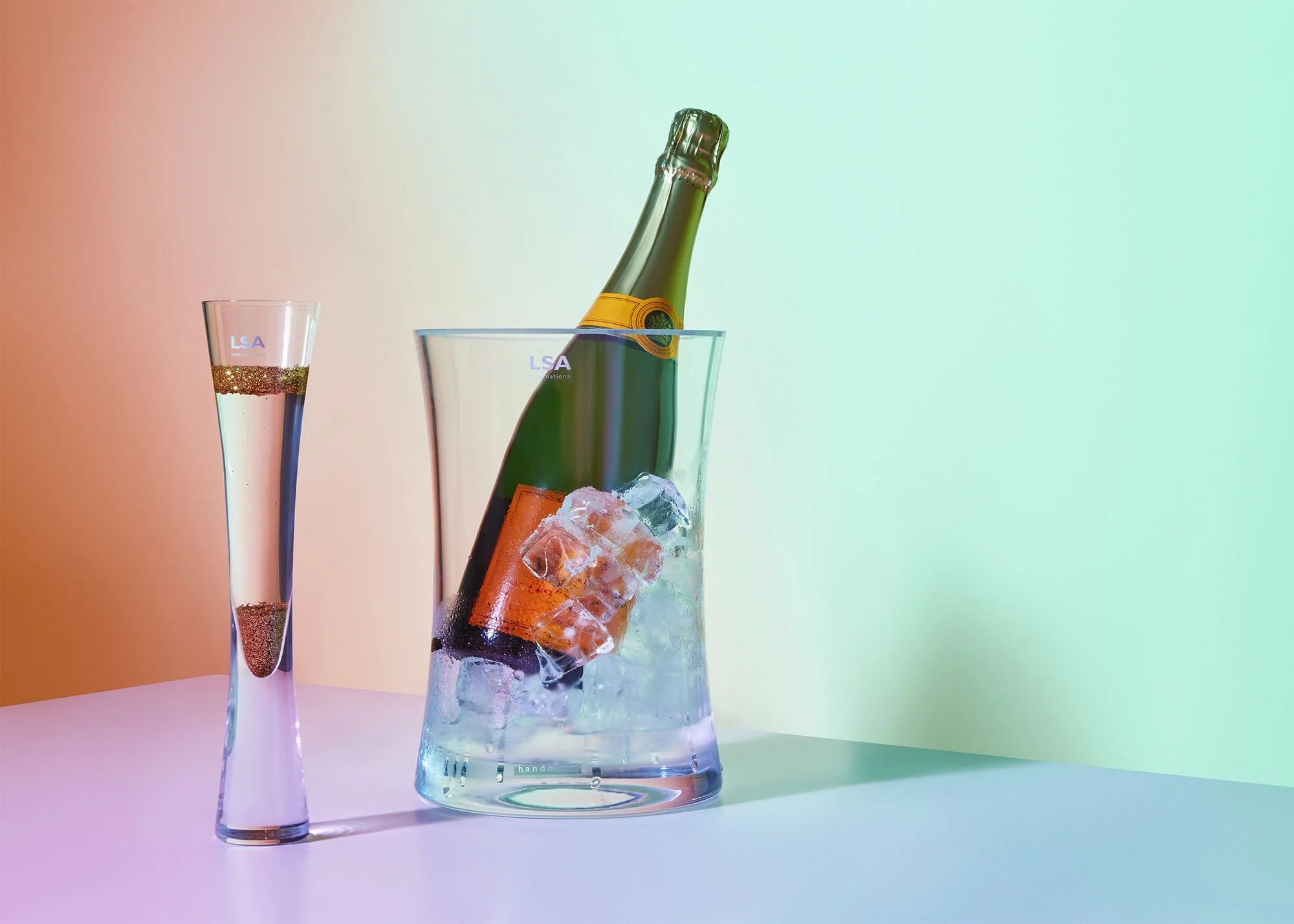 A champagne bottle in an ice bucket and a champagne flute with gold glittered bubbles inside, set against a pastel colored background.